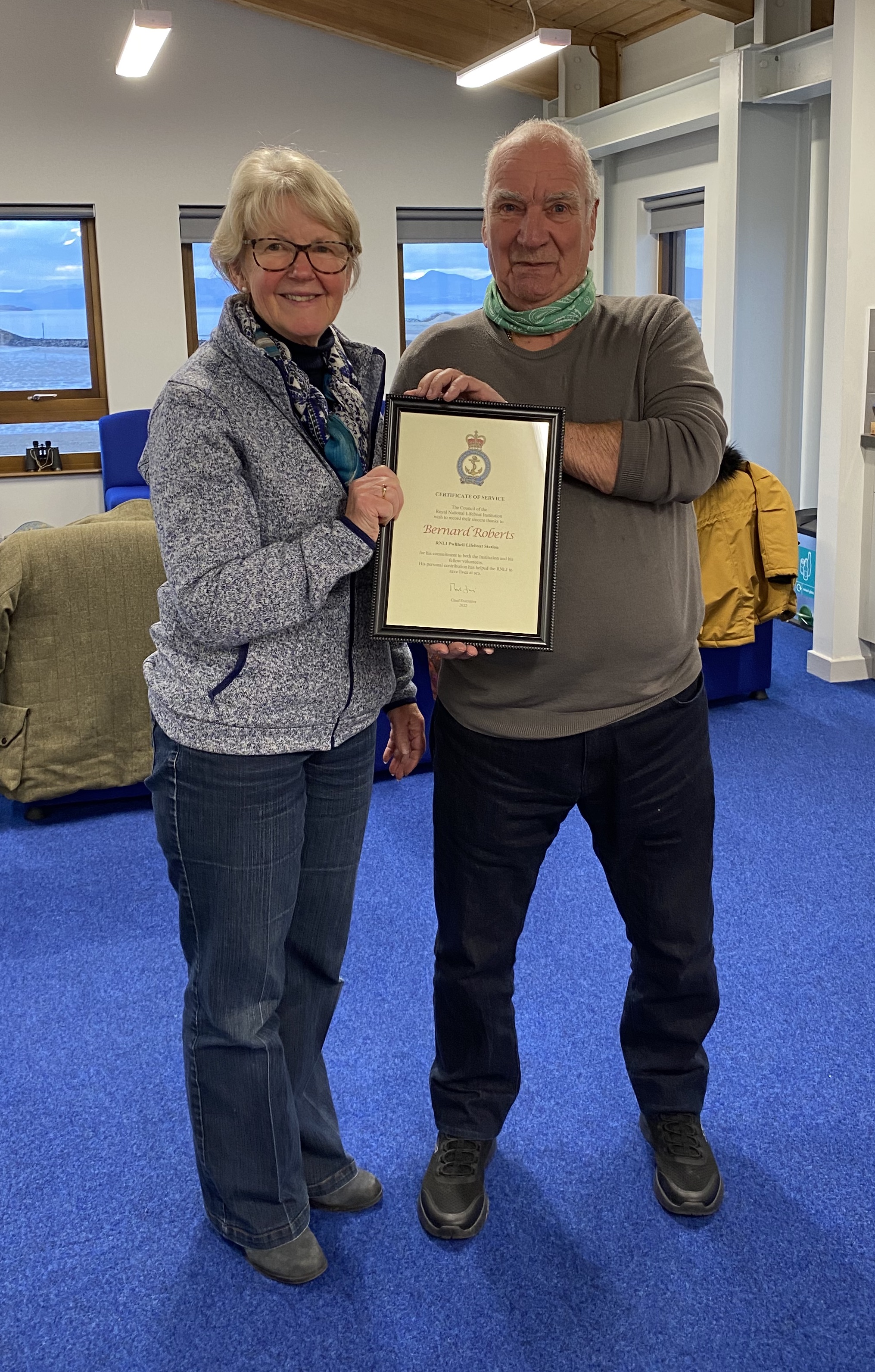 Alison Hayes, Chairperson of Pwllheli RNLI fundraisers stands to the left of Bernard as they both hold the framed RNLI Certificate of Service.
