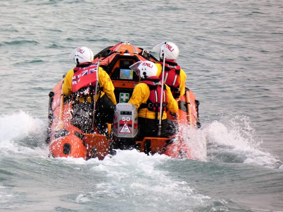 Newquay RNLI D class inshore lifeboat and three crew power away from camera in calm seas