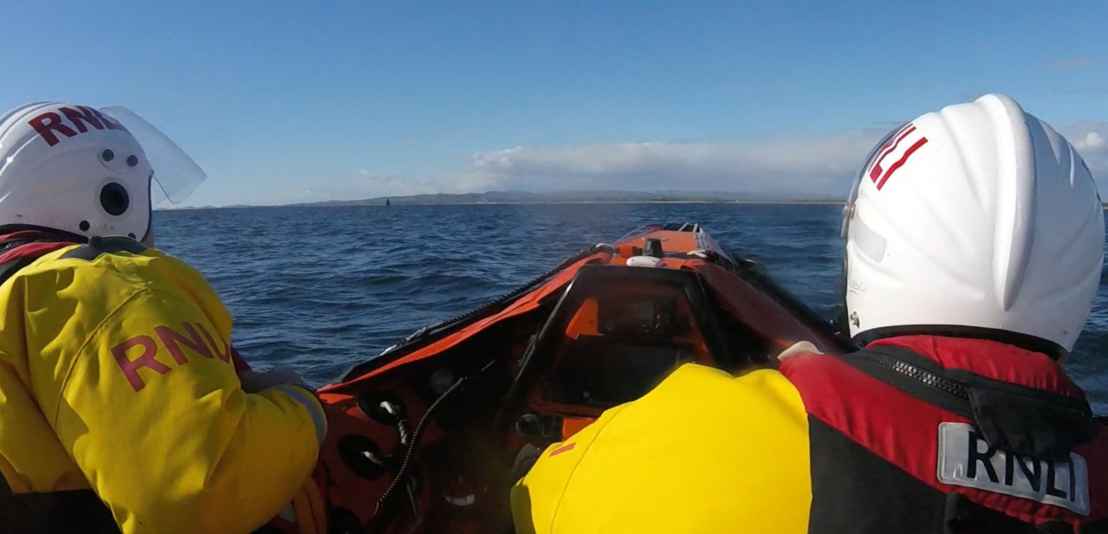 a view from behind two crewmembers in drysuits and helmets as on board the RNLI inshore lifeboat as the look out to sea on a calm day