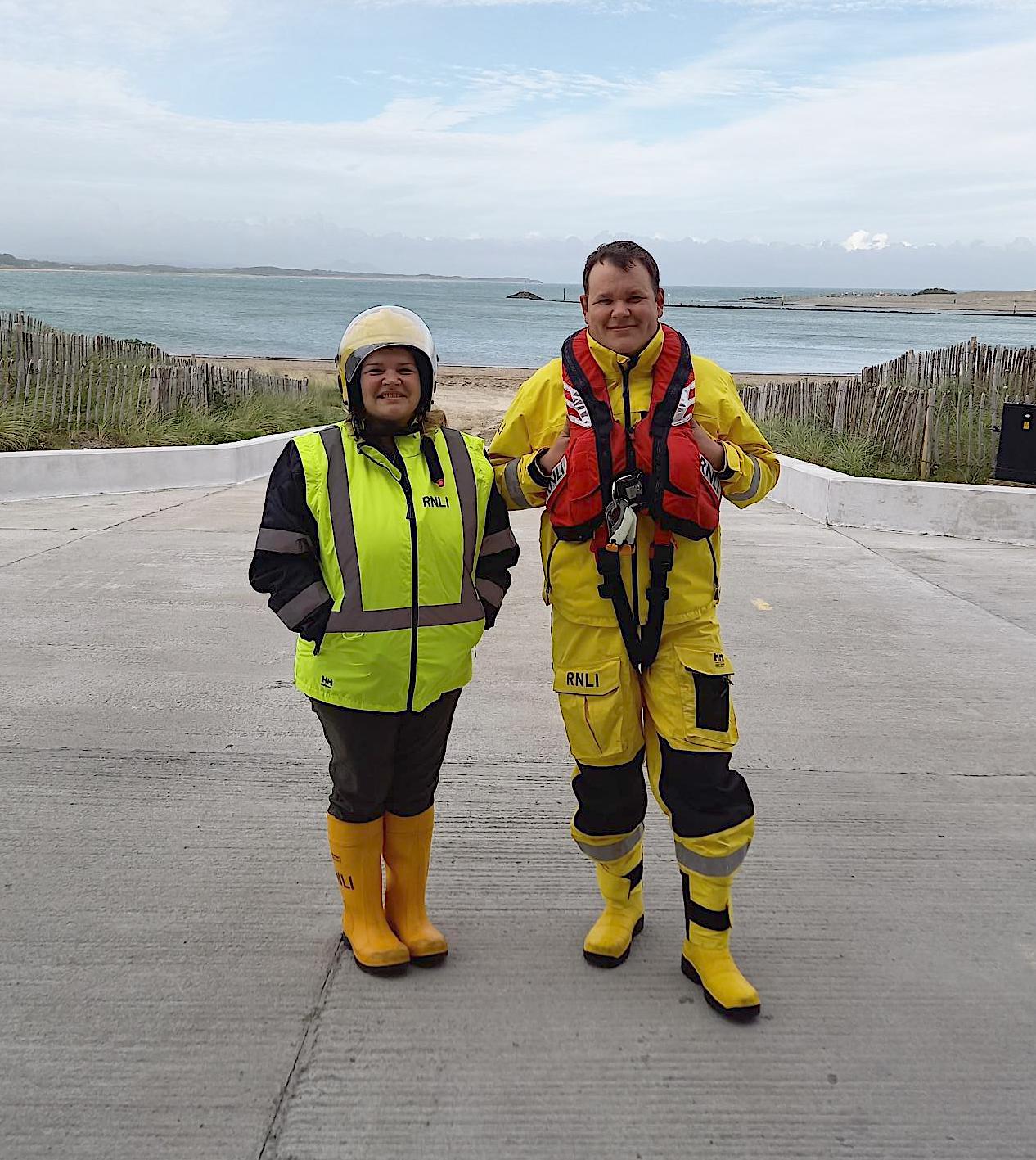 RNLI Pwllheli volunteer crew members Caroline Jones on the left, and Adam Jones standing in their crew kit on the RNLI Pwllheli boathouse slipway with sea behind them