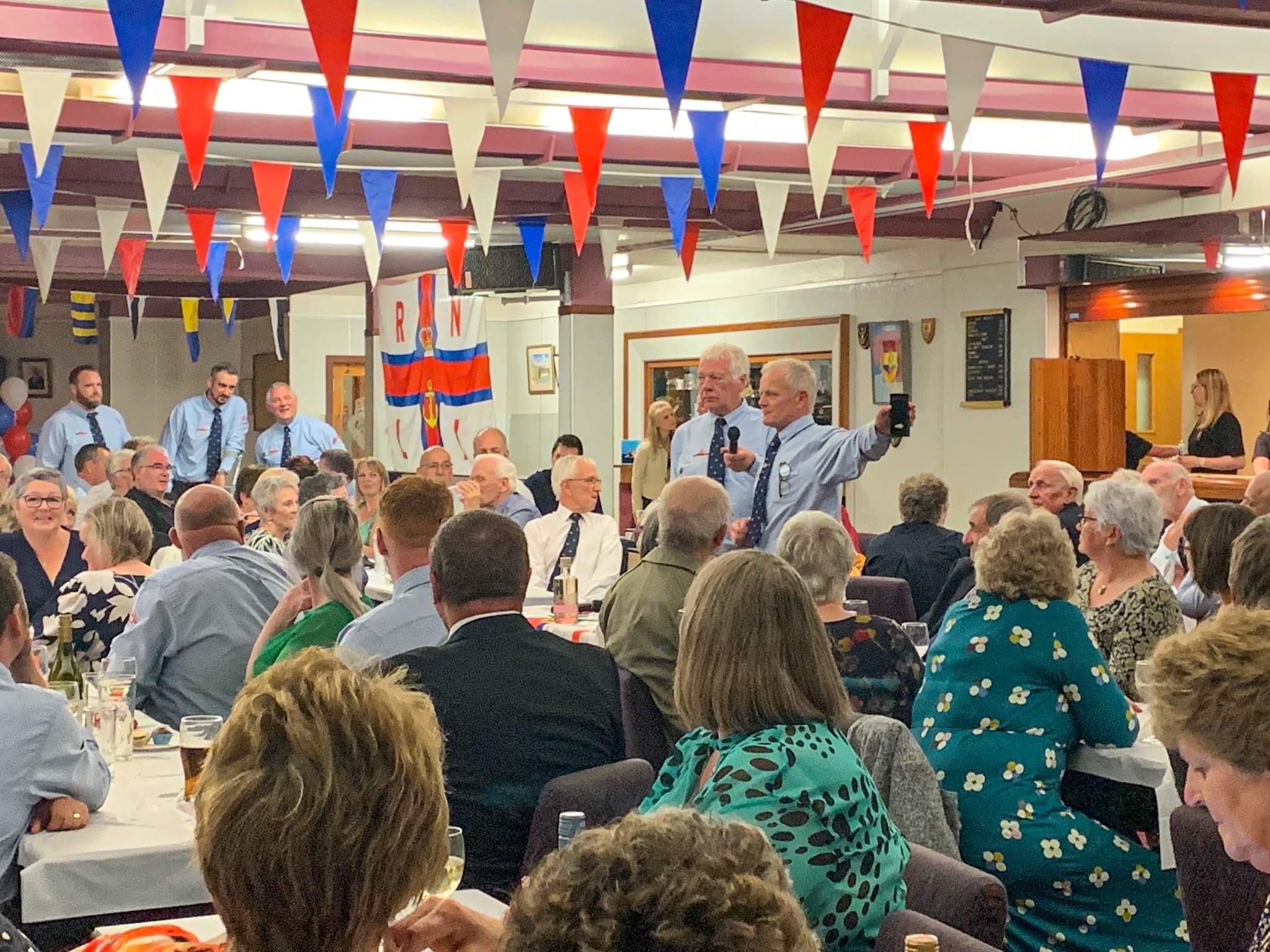 People can be seen seated at tables at the event, with the room decorated with RNLI Flags and bunting.