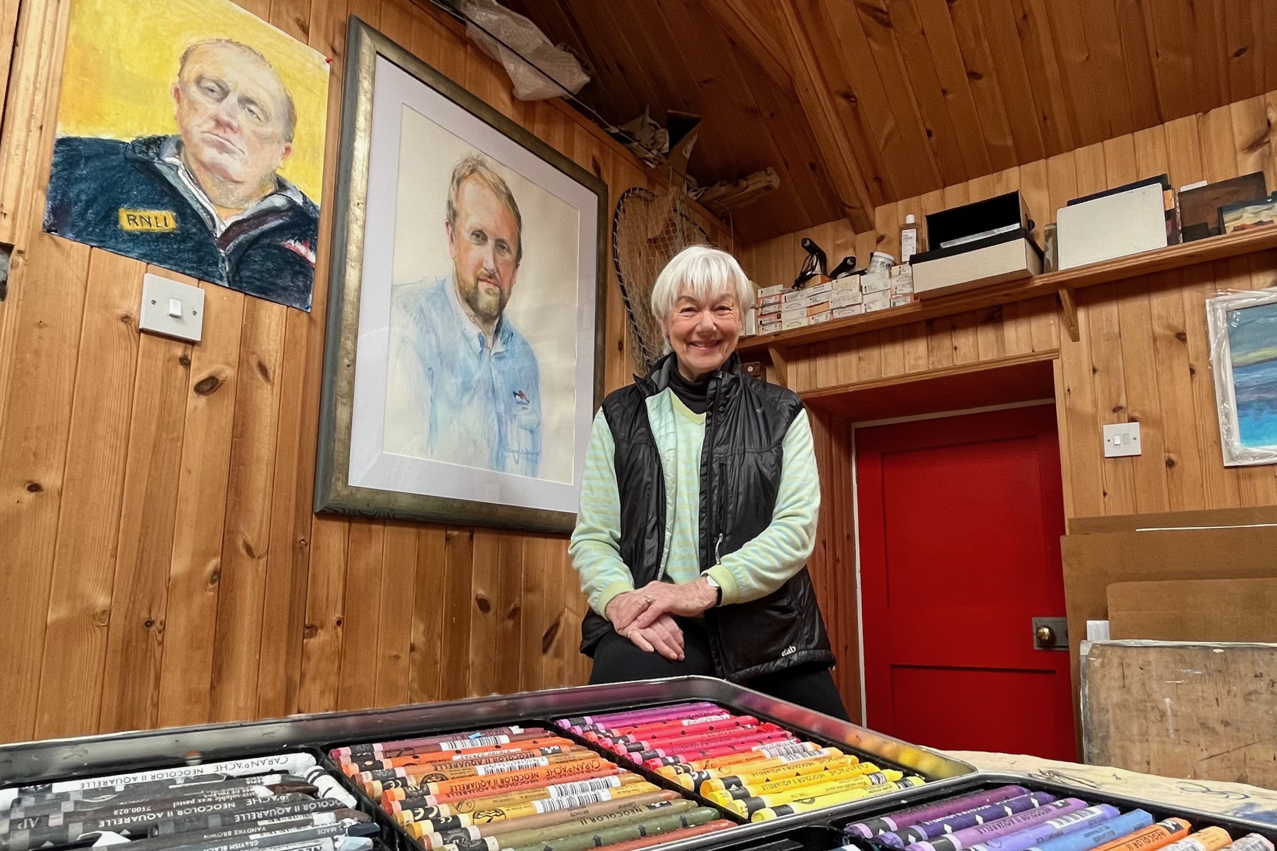 Artist, Heather Dewar, sitting centre on a stool in a wooden clad studio with two portraits hanging on the wall behind. In the foreground is a box os pastels