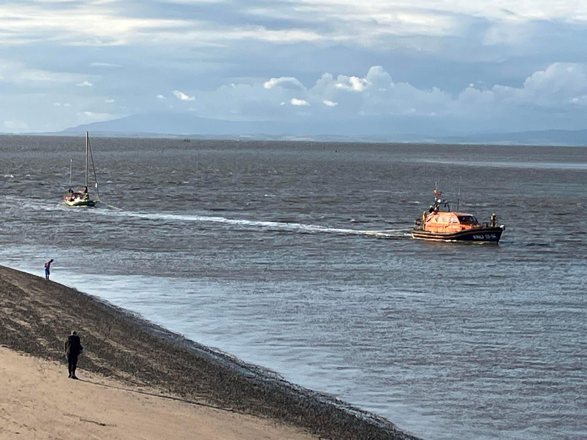 The Shannon class RNLB Kenneth James Pierpoint pulling a twin-masted, green-hulled yacht on a tow line. In the foreground we see a section of Fleetwood beach where two people are watching the boats come in.