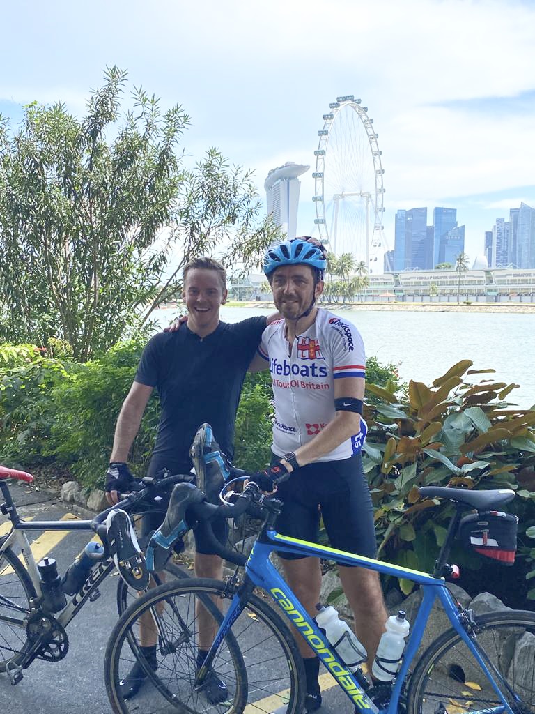 A man wearing cycling gear and with a bike stands beside a friend on the banks of the river Thames with the London Eye behind him