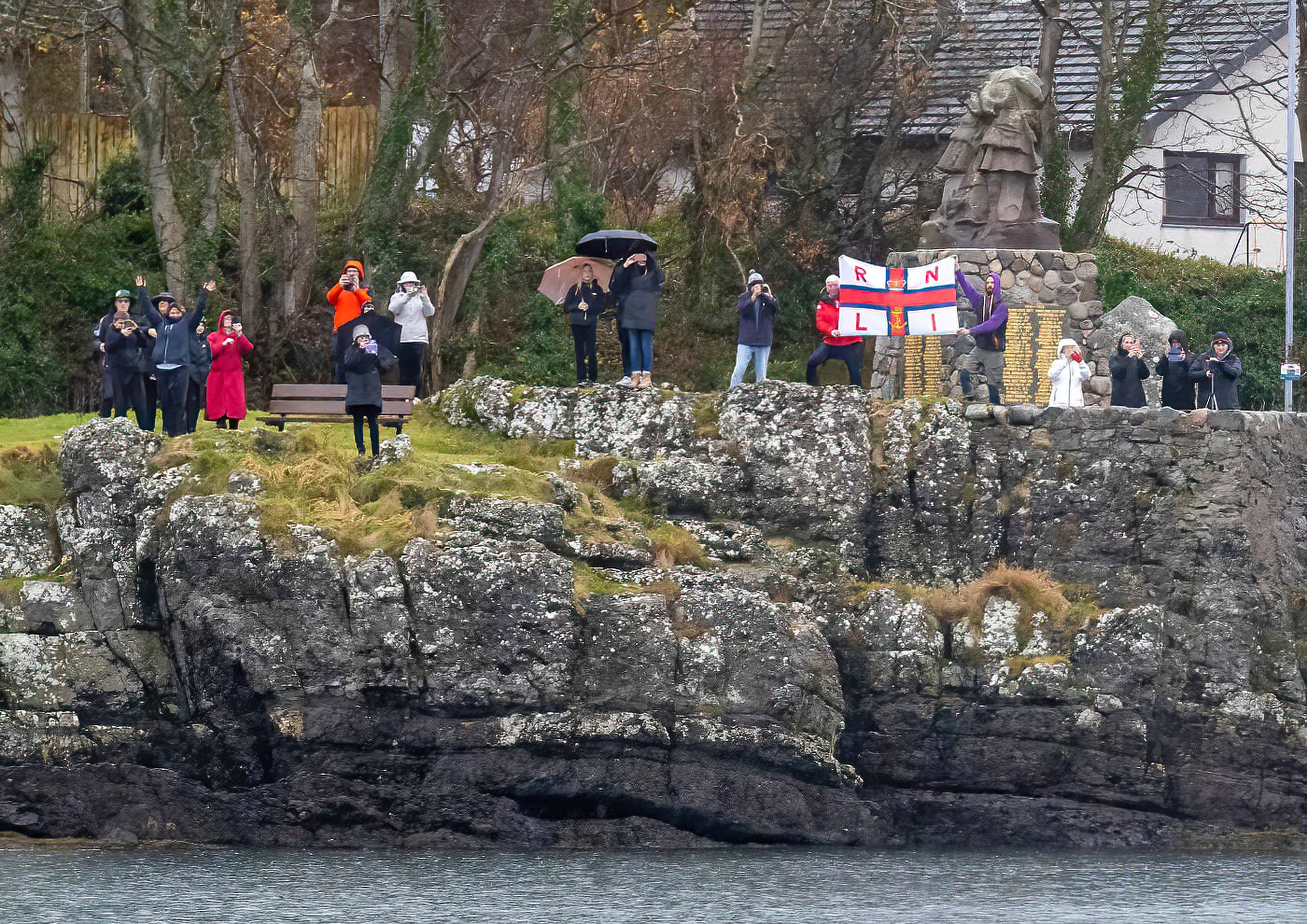 Photo from the water of people standing in the rain by Oban's war memorial