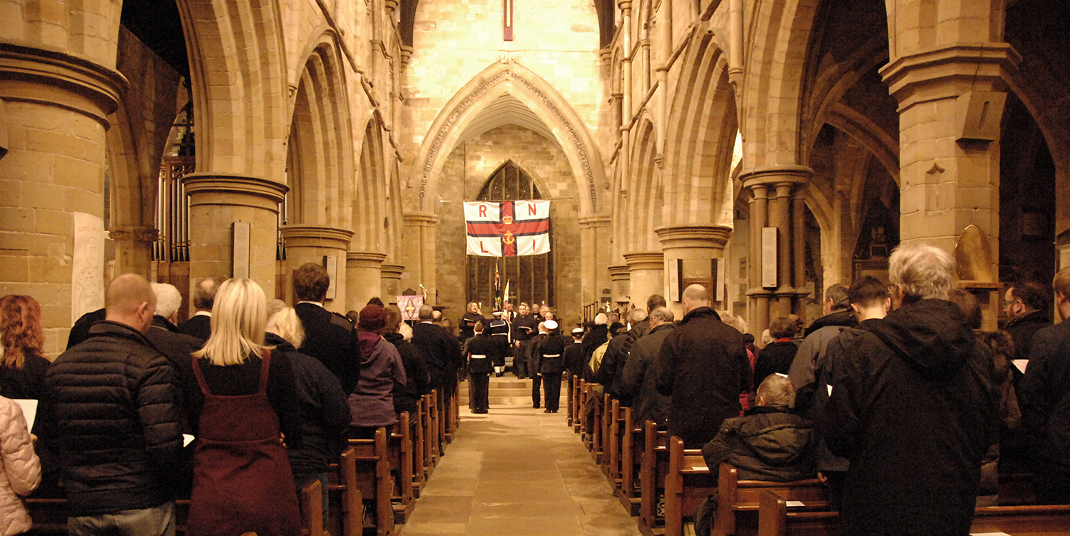 View of the congregation at Saint Mary's Church, Scarborough during the Scarborough RNLI ECJR memorial service. In the centre of the image is the RNLI ensign, and below that in the distance is the standard bearers and Filey Fisherman's Choir.To the left and right of the aisle is the congregation who are wearing black or dark coloured clothing, sat on dark wood benches. They are surrounded by the stone pillars and aches of the church's architecture..