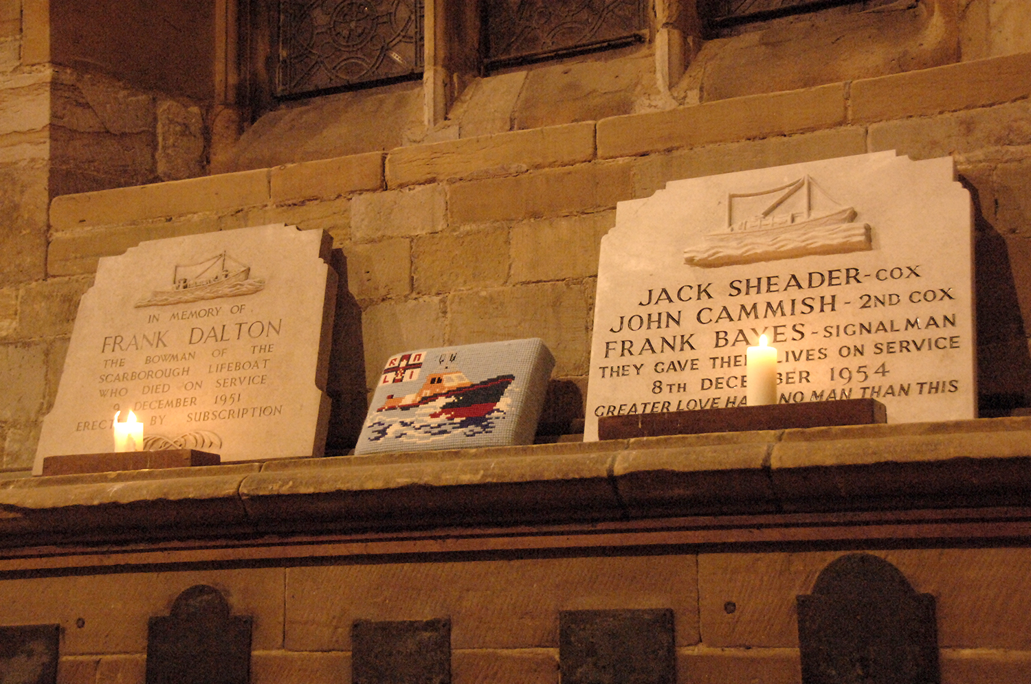 The left hand marble memorial has inscribed 'In memory of Frank Dalton the bowman of the Scarborough Lifeboat who died on service on 9 December 1951 erected by subscription'. In the centre is an embroidery of an all-weather lifeboat with the RNLI ensign to the left of the vessel. The right hand marble memorial has inscribed 'Jack Sheader- Cox, John Cammish - 2nd cox, Frank Bayes - signalman. They gave their lives on service 8th December 1954. Greater love hath no man than this.'