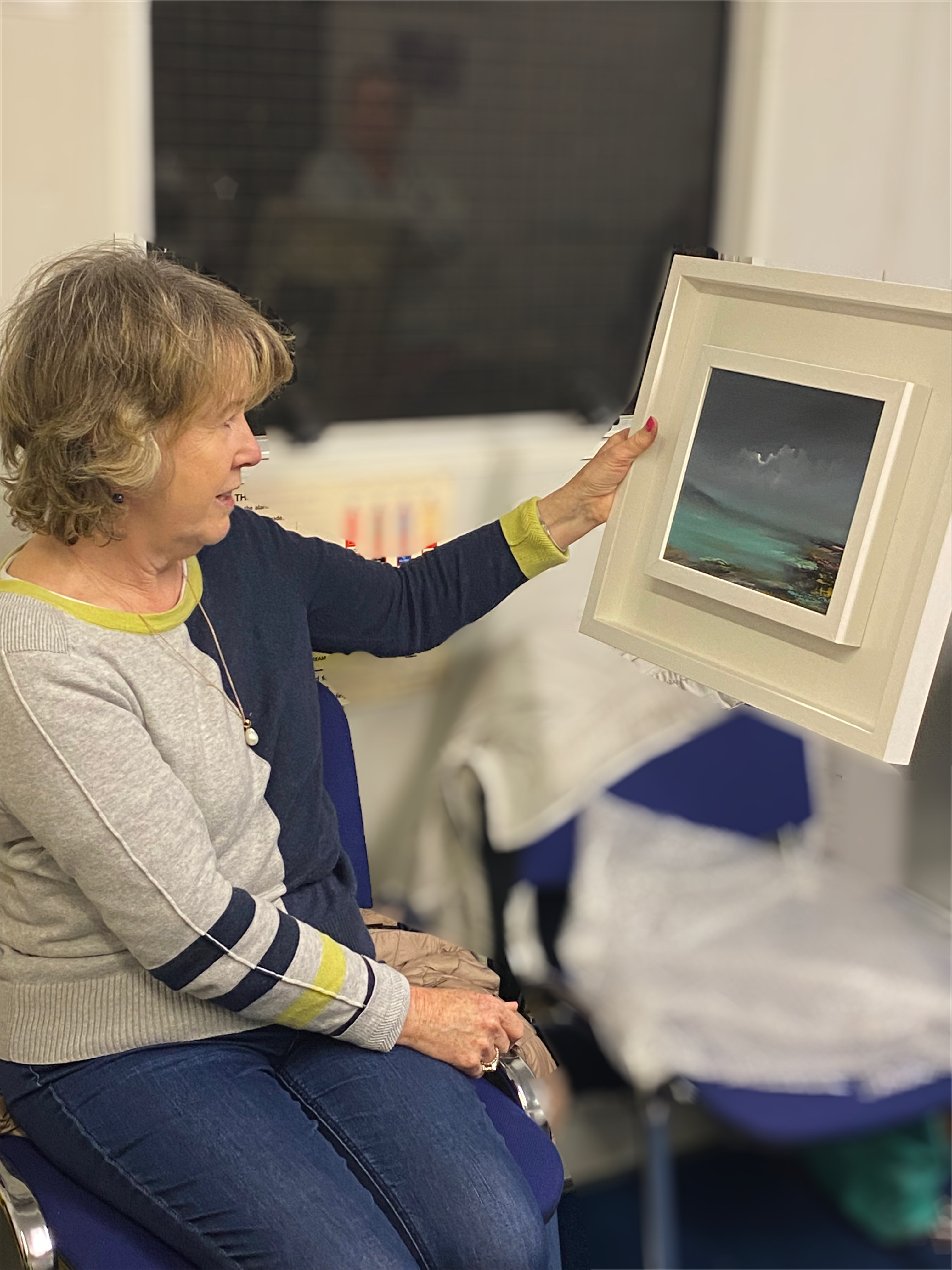 Catherine is seated and looking at the oil painting of a moody sky over water