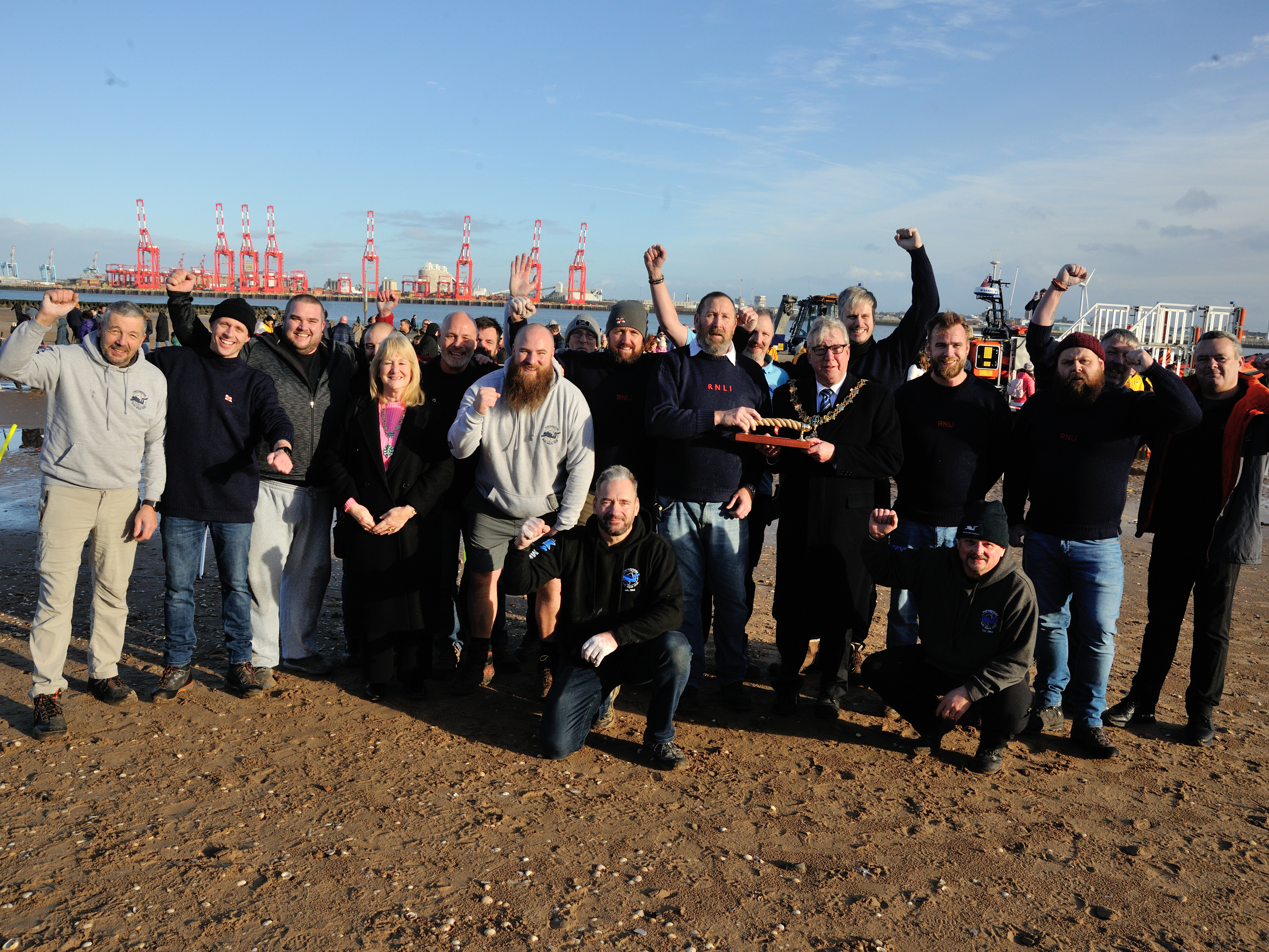 New Brighton RNLI's volunteer tug of war team pose on the beach alongside their opponents from Mersey Diver's Club. The Mayor presents the trophy to one of the RNLI crew. Fists and arms are raised in good-spirited celebration. Striking red cranes are visible across the river in the background. 