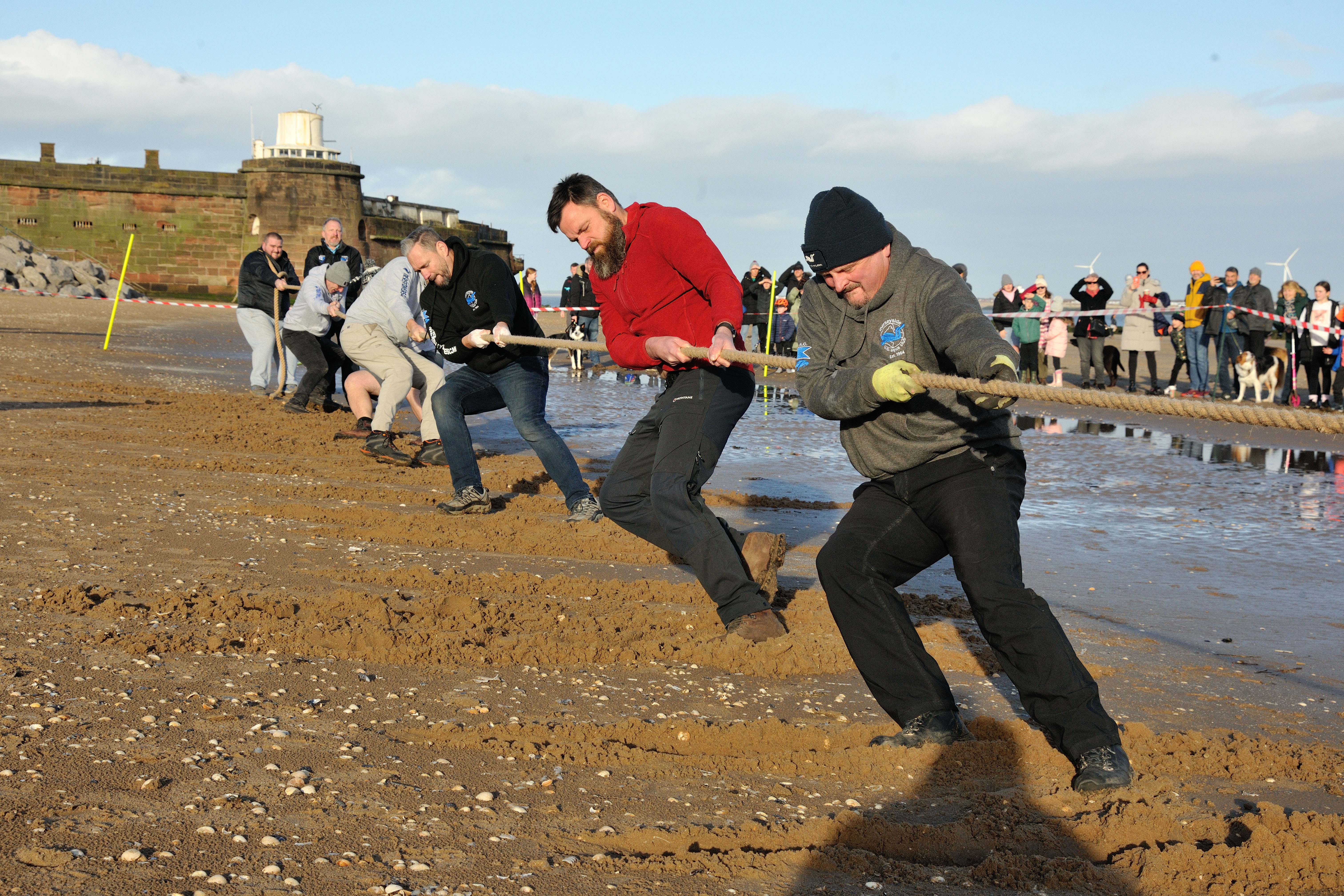 The tug of war team from Mersey Diver's Club, all kitted out for the winter cold in varied attire, strain and lean back, putting all their effort into pulling on the rope. In the midground, crowds watch on, in the background Fort Perch Rock is visible.
