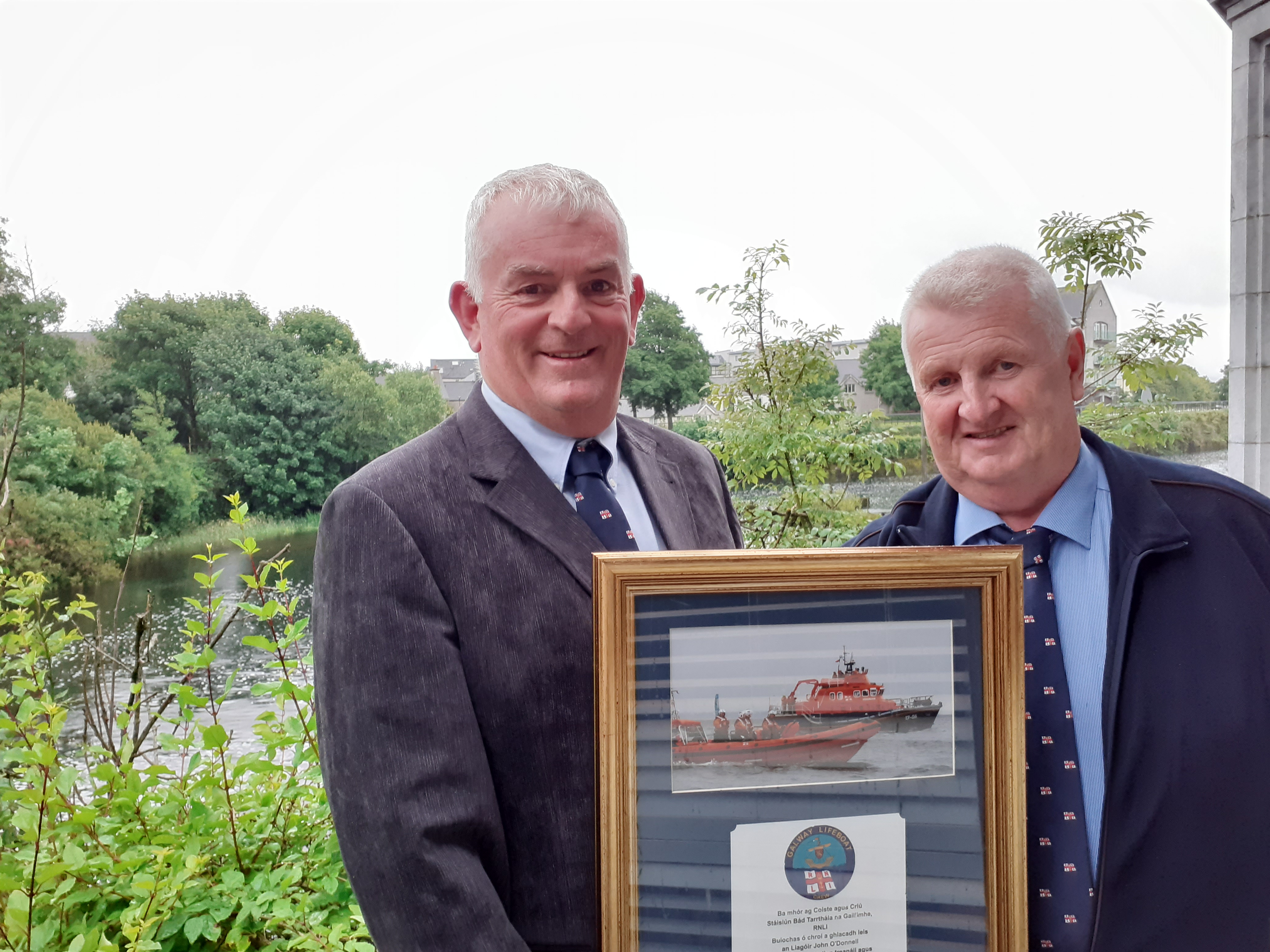 Mike Swan from Galway RNLI holding a large framed photo of two lifeboats with a dedication underneath, standing beside John O'Donnell retired Coxswain from Aran Islands RNLI. In the background, trees and a glimpse of the River Corrib between the foliage