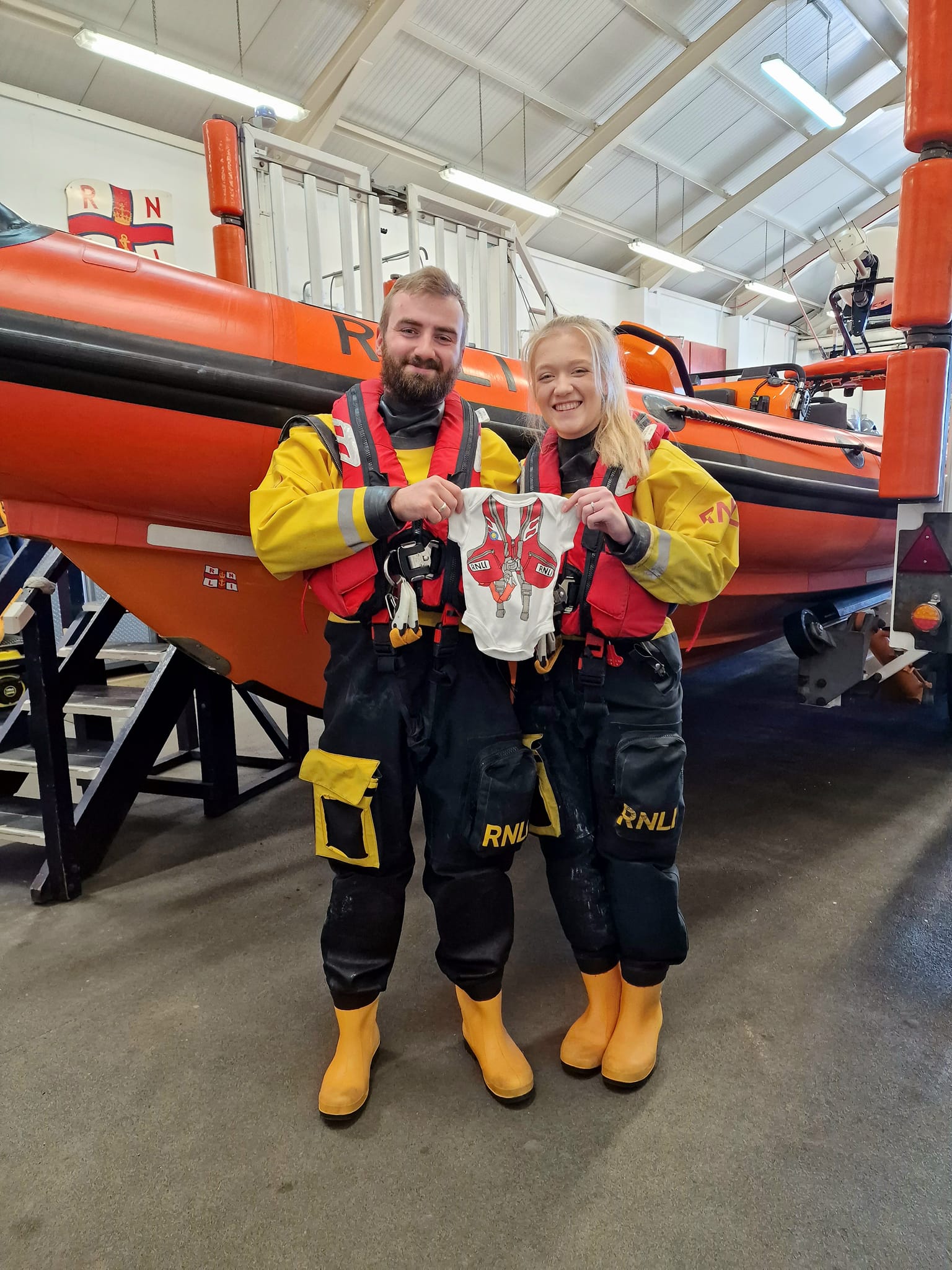 Tom and Emily stand in their dry suits in front of New Brighton RNLI's Atlantic-85 lifeboat. They hold up a small baby grow with a lifejacket image printed on it.
