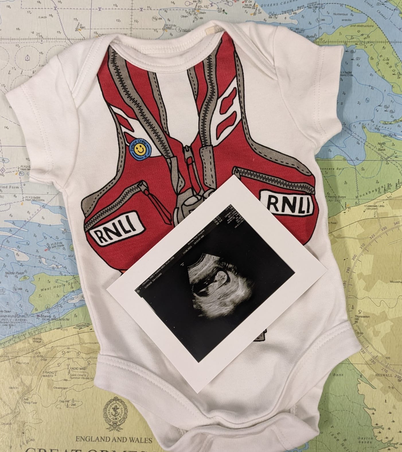 A small baby grow featuring a print of a RNLI lifejacket sits on an admiralty chart. On top is an initial pregnancy scan photo.
