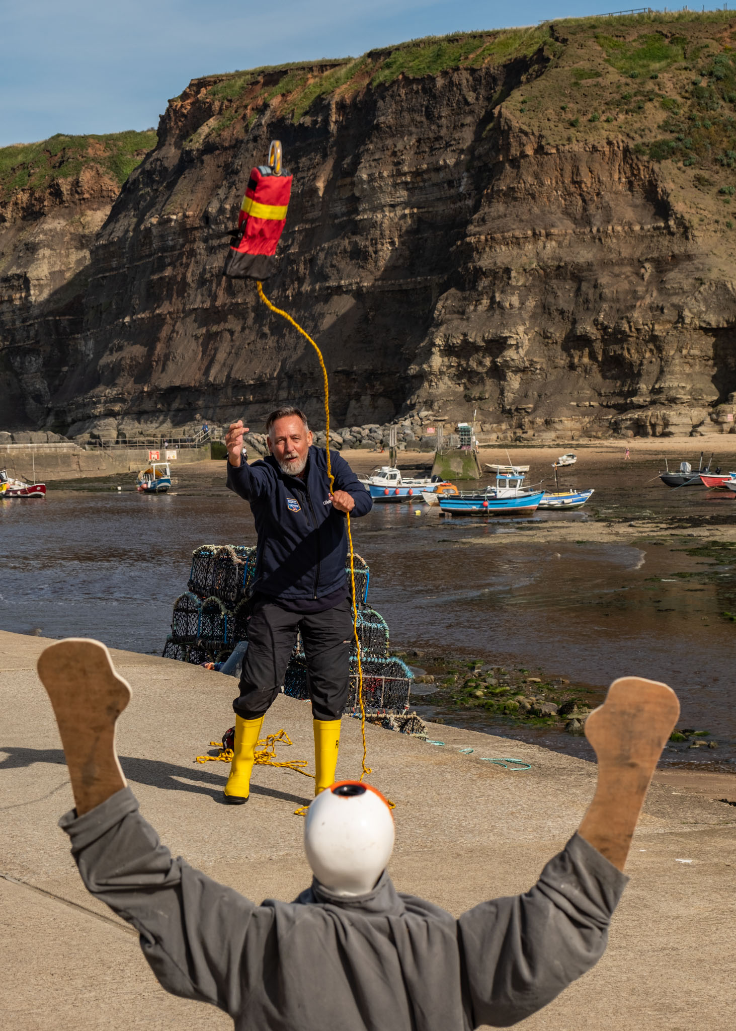 A man, wearing yellow RNLI wellies, throws a red throw bag toward a mannequin with it's arms in the air. The throw bag is in mid air and is heading straight for the mannequin. There is a harbour and boats and a cliff face in the background.