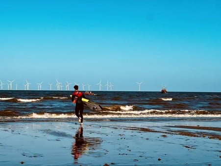 A lifeguard heads out to work in rolling waves at the beach with a wind farm in the distance