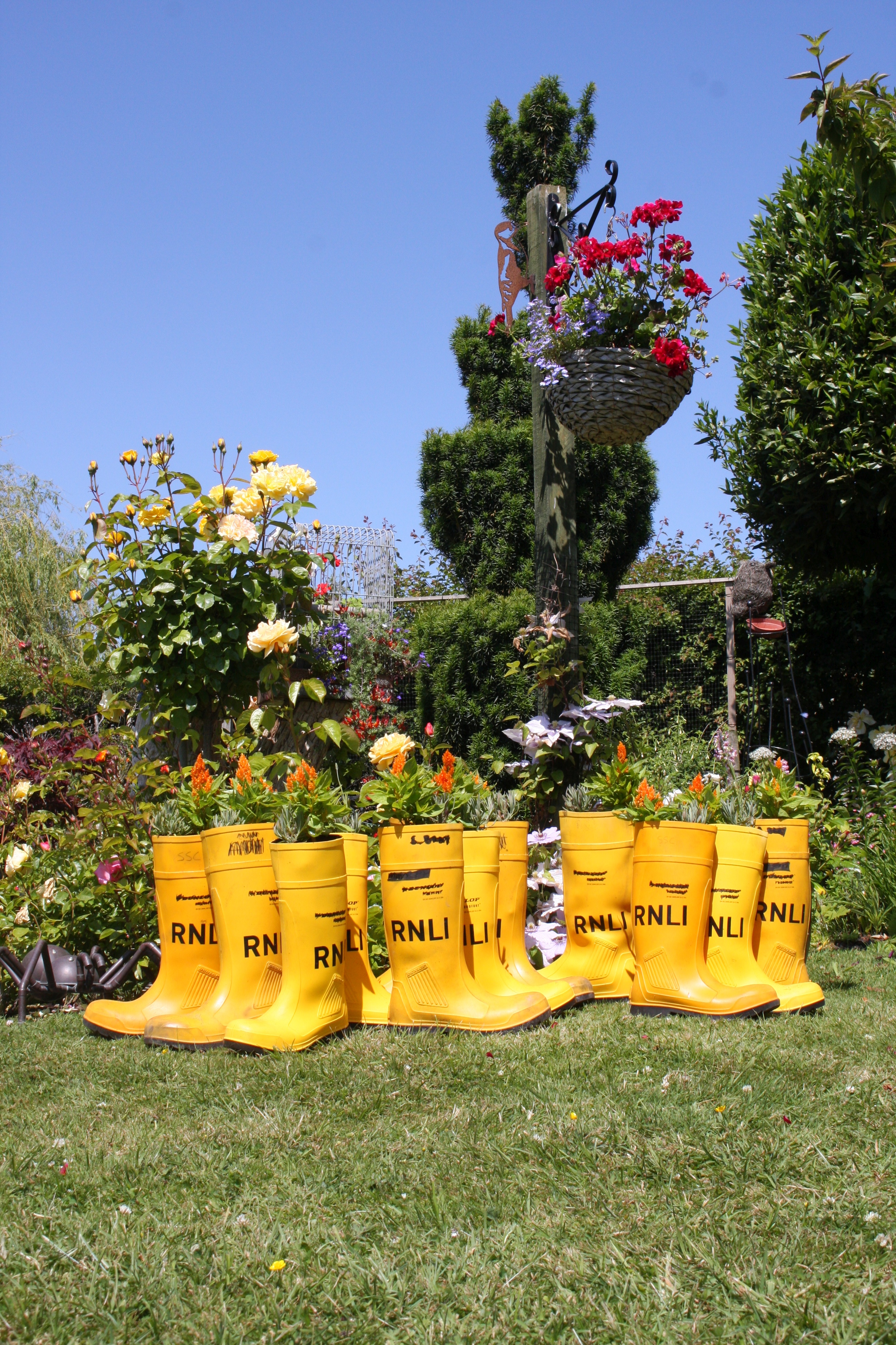 Yellow lifeboat wellies with plants in them sit on a green lawn with a blue sky above.