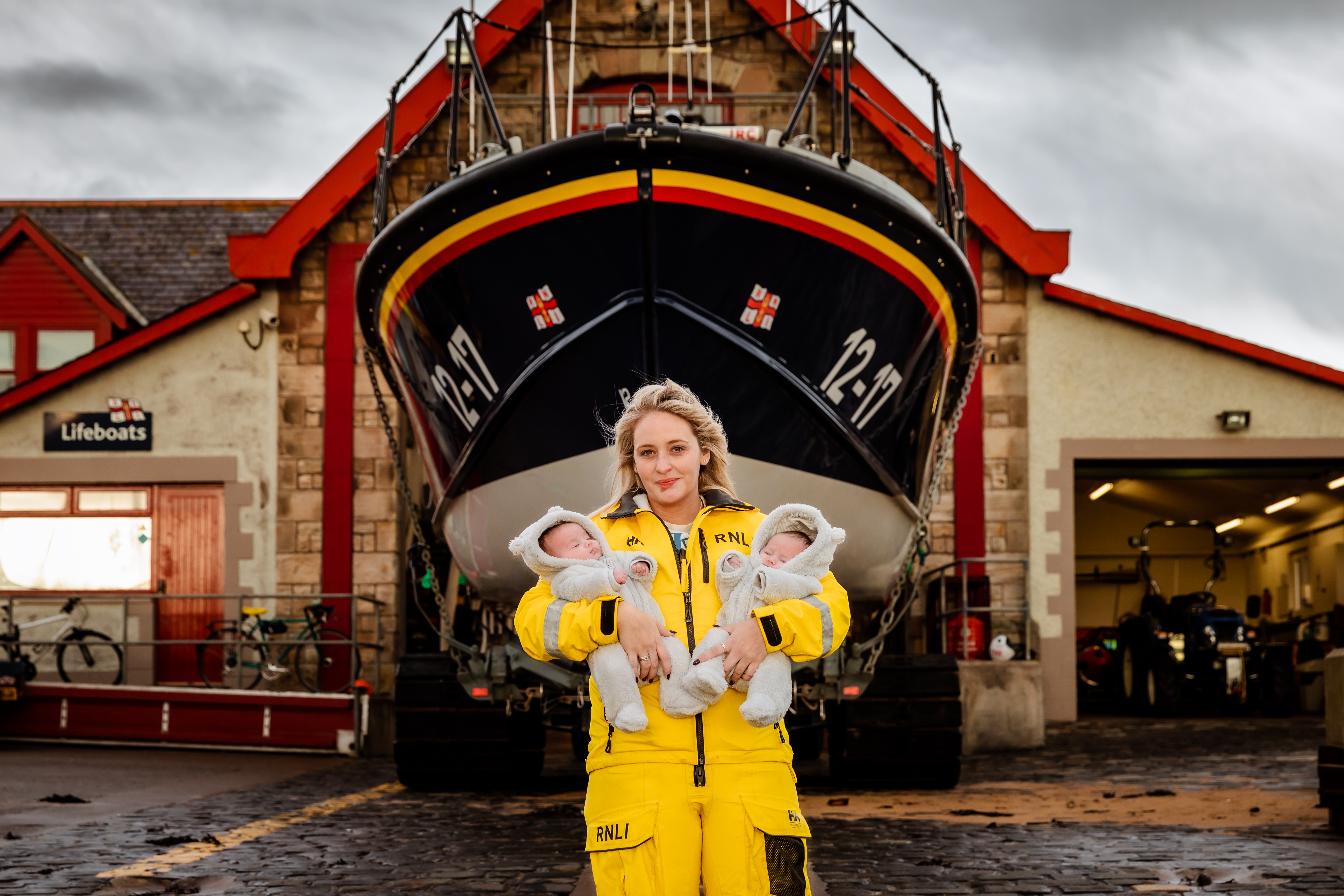 Female volunteer in yellow RNLI kit holds baby girls in front of Mersey class lifeboat.