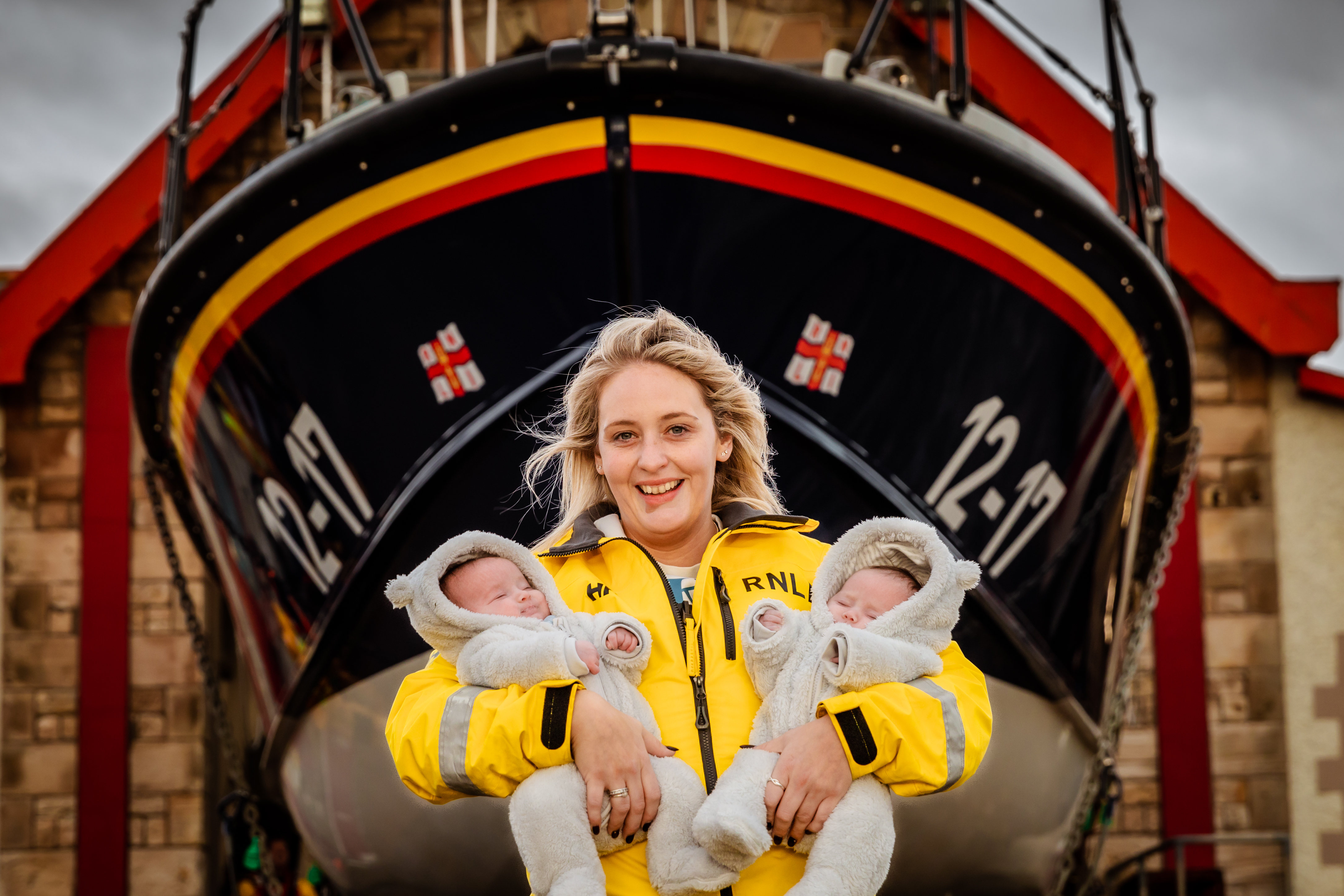 Female volunteer in yellow RNLI kit holds baby girls in front of Mersey class lifeboat.