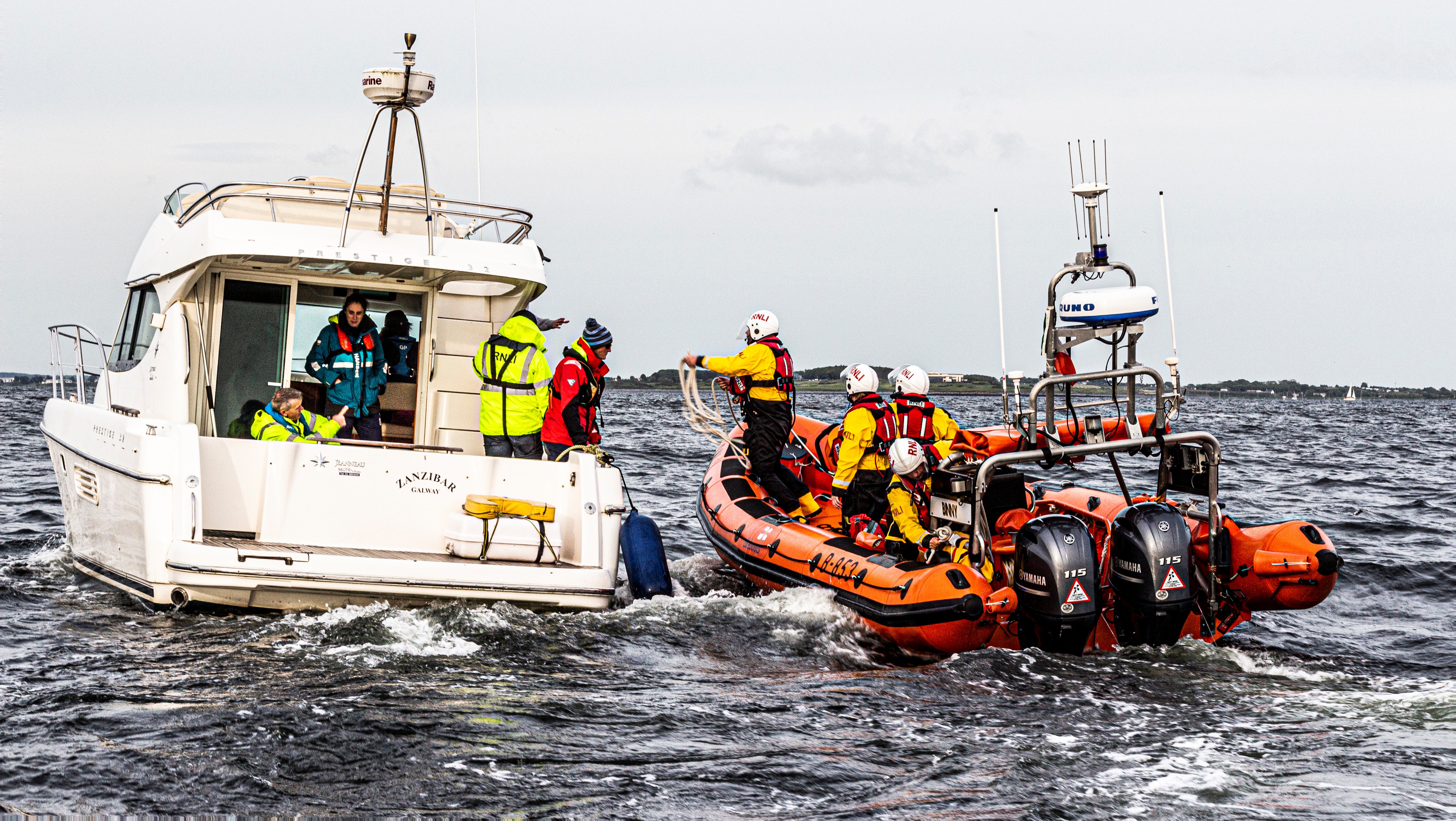 Inshore lifeboat with four crew coming alongside a cruiser on the water in Galway Bay. A RNLI crew member is preparing to throw a line to one of the crew on the cruiser