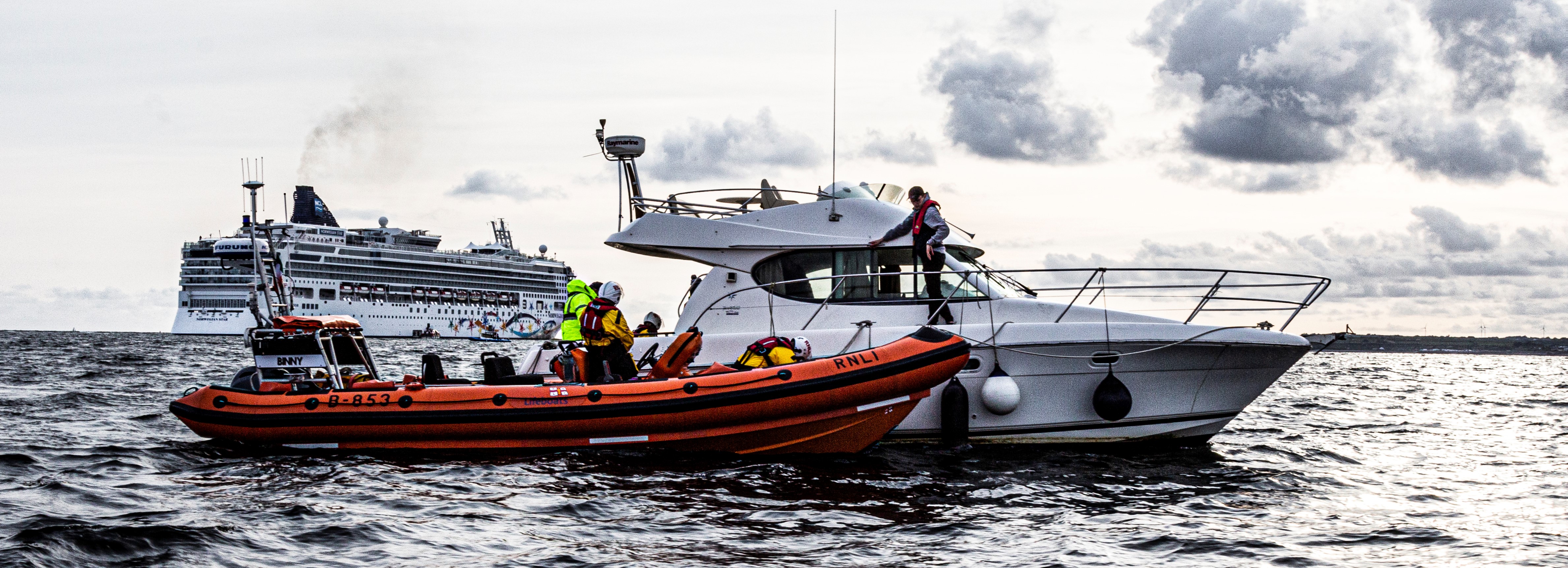 The inshore lifeboat alongside a cruiser as part of the trauma simulation exercise with two RNLI crew members visible. There is one crew member from the cruiser on the deck of the cruiser. In the background there is a very large cruise ship which was departing Galway after a short visit.