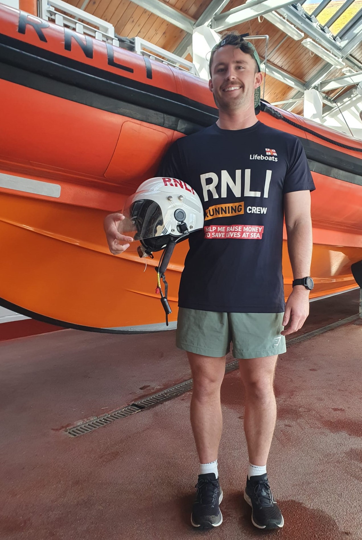 Mudeford RNLI volunteer Scott Harburn in running gear holding an RNLI helmet standing next to an orange Atlantic 85 lifeboat