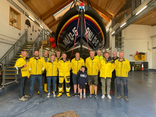 Members of the 2023 Scarborough RNLI crew al wearing yellow RNLI jackets stood with Seb, who is wearing his navy RNLI hoody, in front of the all-weather lifeboat 'Frederick William Plaxton'