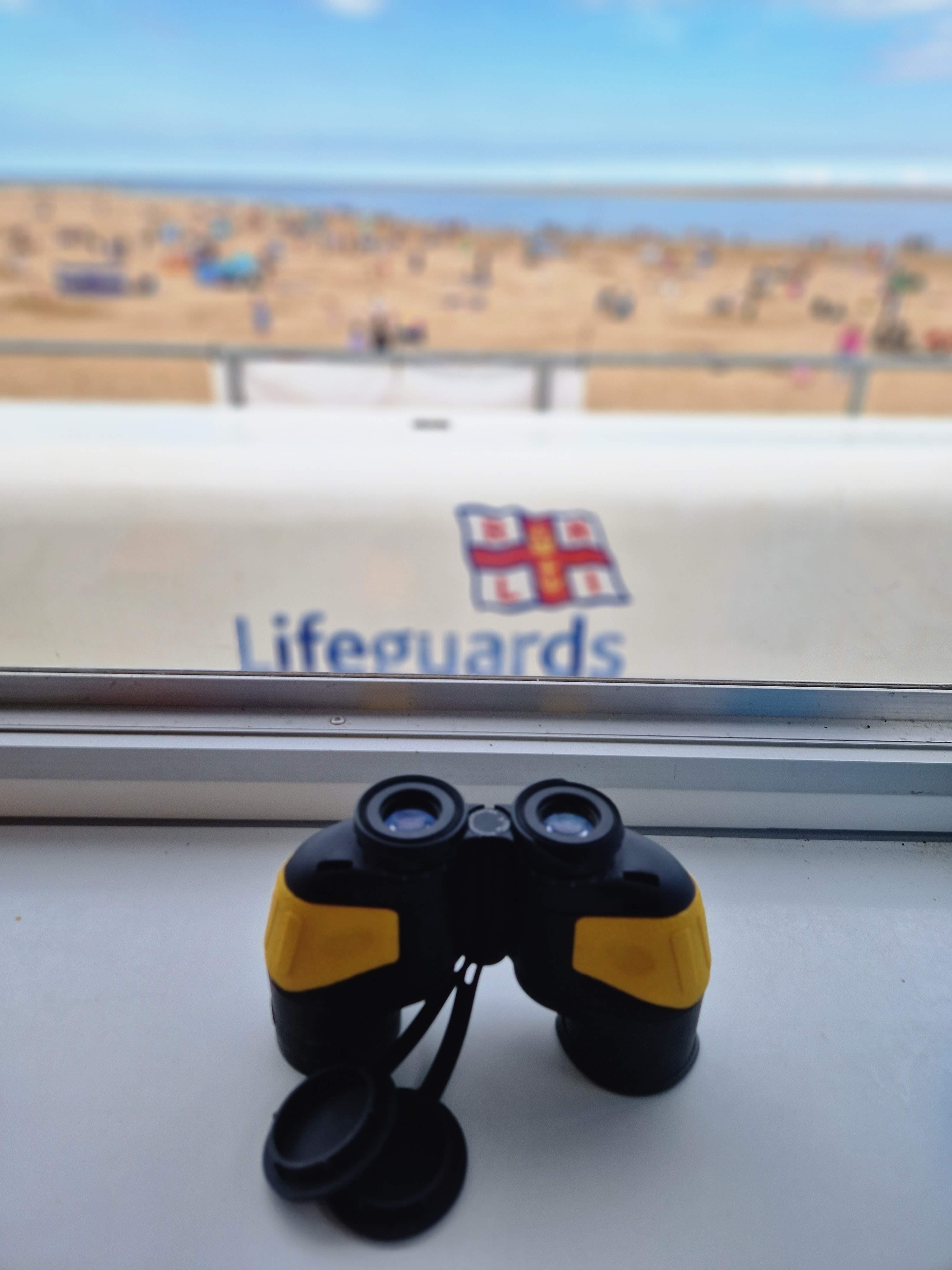 A photo of a pair of binoculars used by the lifeguards and the RNLI lifeguards emblem in the background and the beach and swimmers in the background