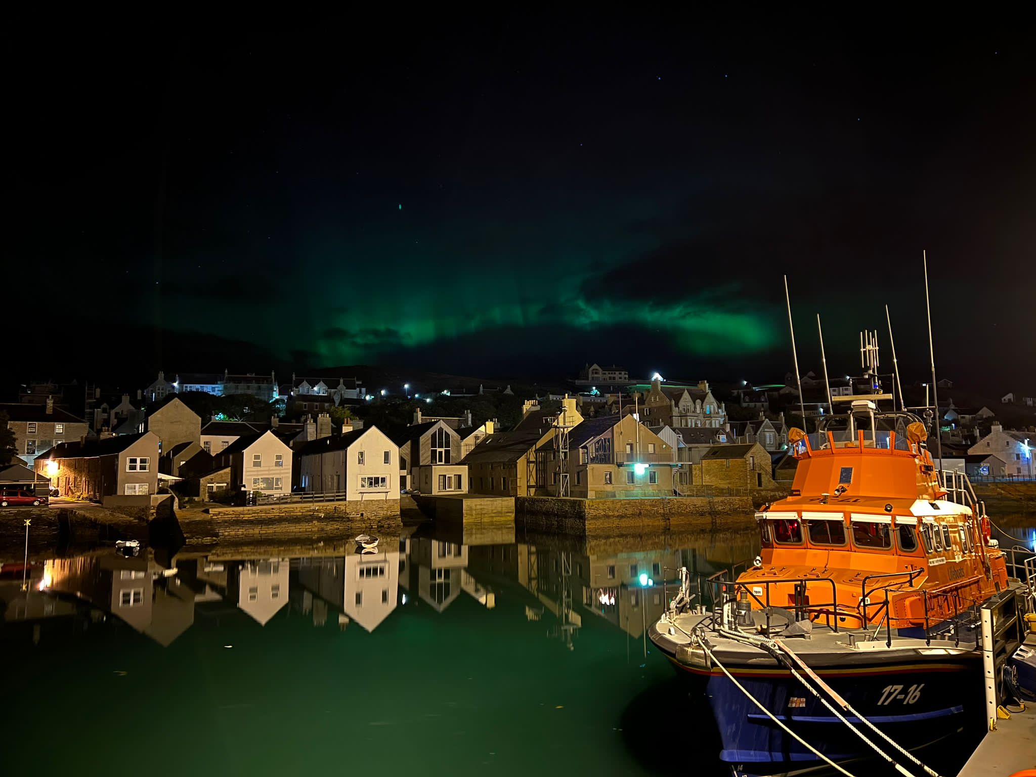 Stromness Lifeboat is shown on her mooring (right) with the town behind and a green smudge of northern lights in the sky above