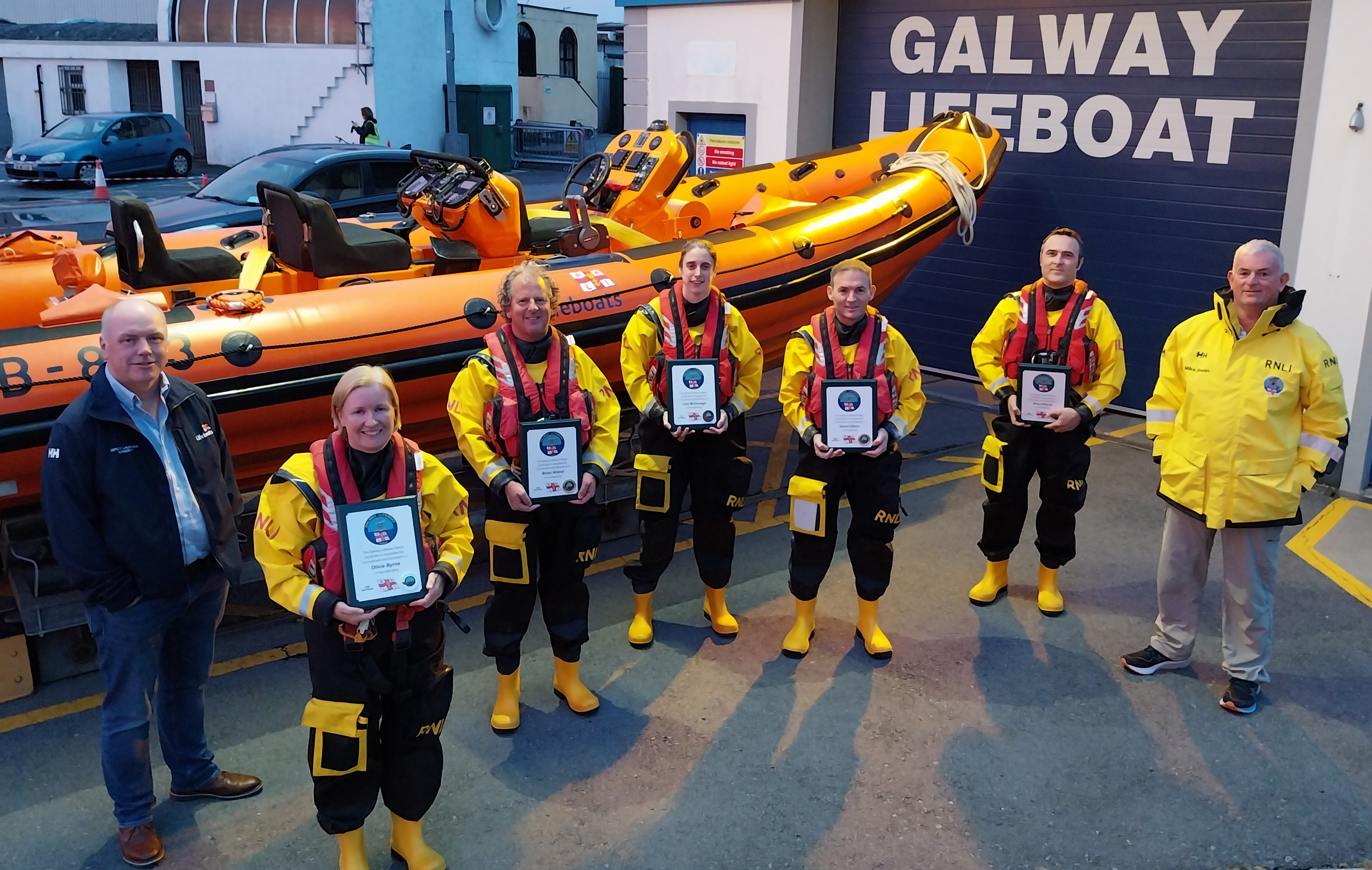 Seven crew standing outside Galway Lifeboat Station with the Atlantic 85 inshore lifeboat in the background (on shore). Five of the crew are wearing dry suits and lifejackets and are holding their long service awards which are framed certificates. It is evening with low light.