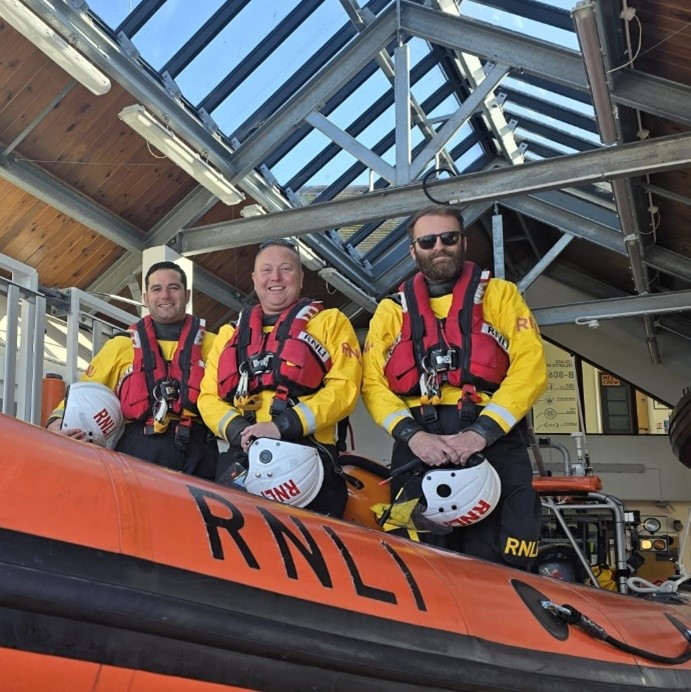 Three men in RNLI drysuits aboard an orange RNLI Atlantic 85 rib