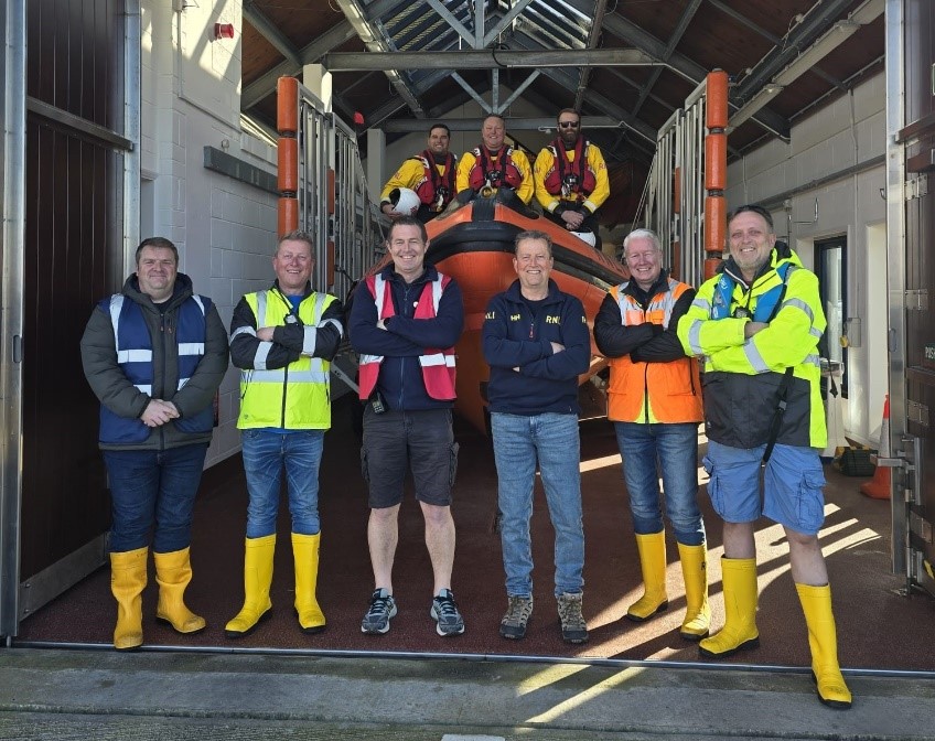 A group of RNLI volunteers stand in front of an orange RNLI Atlantic 85 rib with three crewmembers onboard.