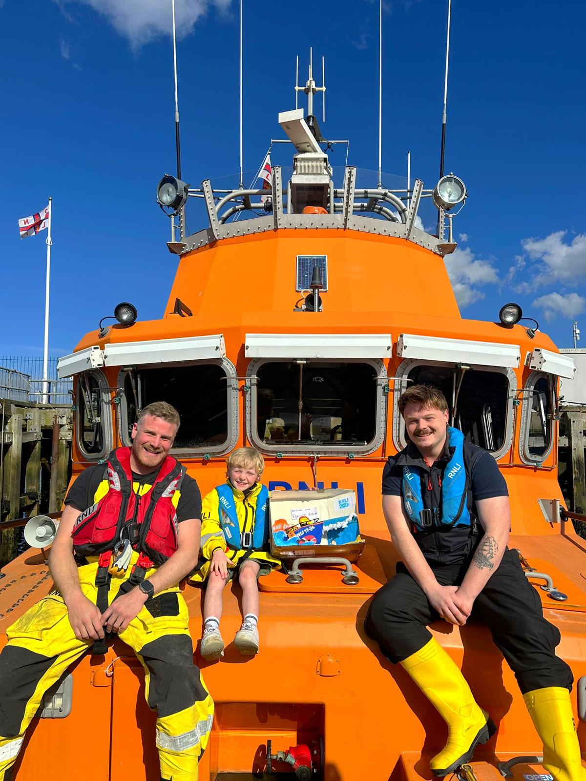 Two crew members either side of boy and his Easter egg RNLI creation. Sitting on the front of Severn Class Lifeboat.