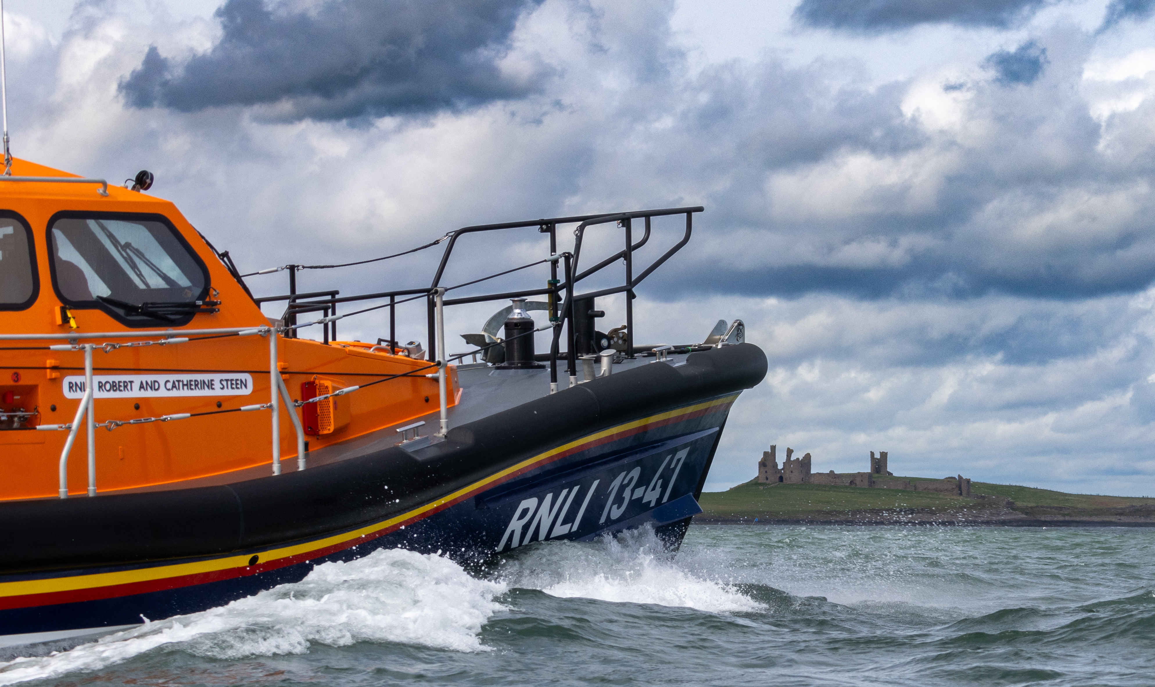 The bow of Shannon Class lifeboat at sea, in the background Dunstanburgh Castle