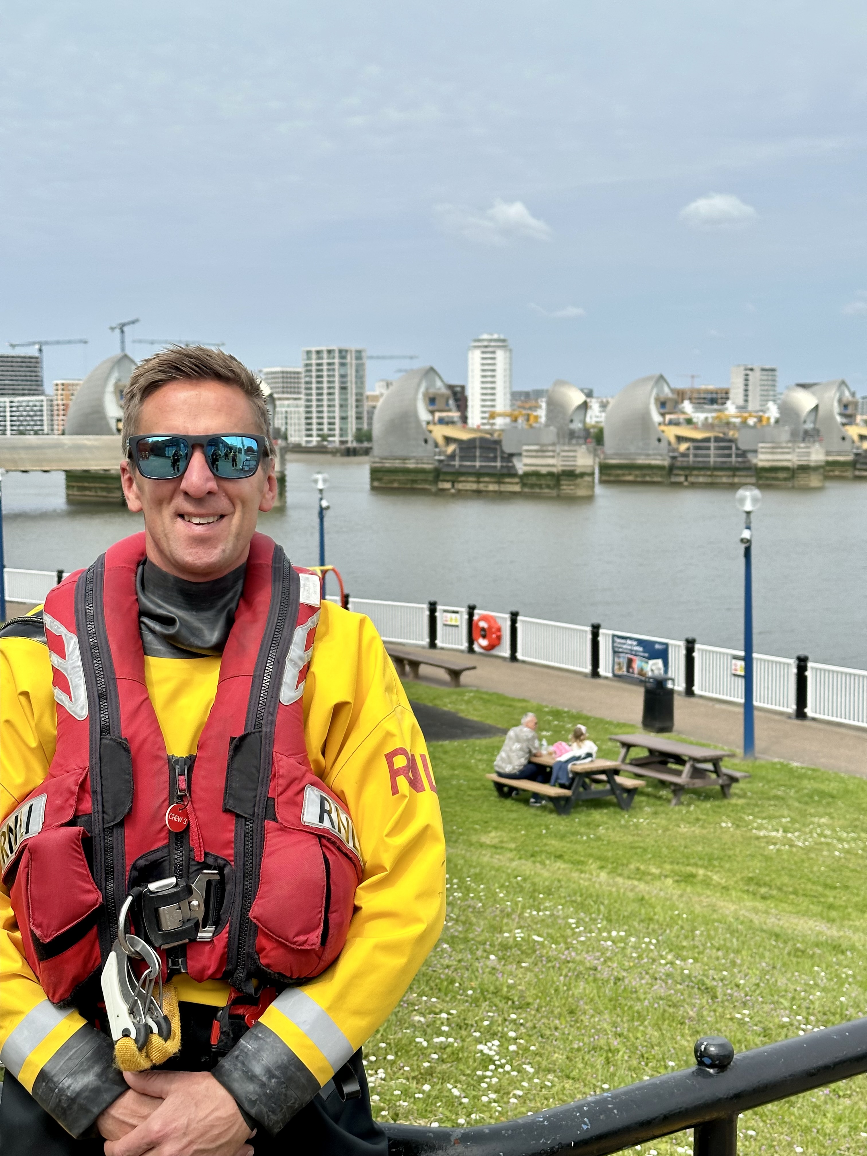 A man stood in front of the River Thames, Thames Barrier, in inshore lifeboat crew kit including yellow wetsuit and red lifejacket.