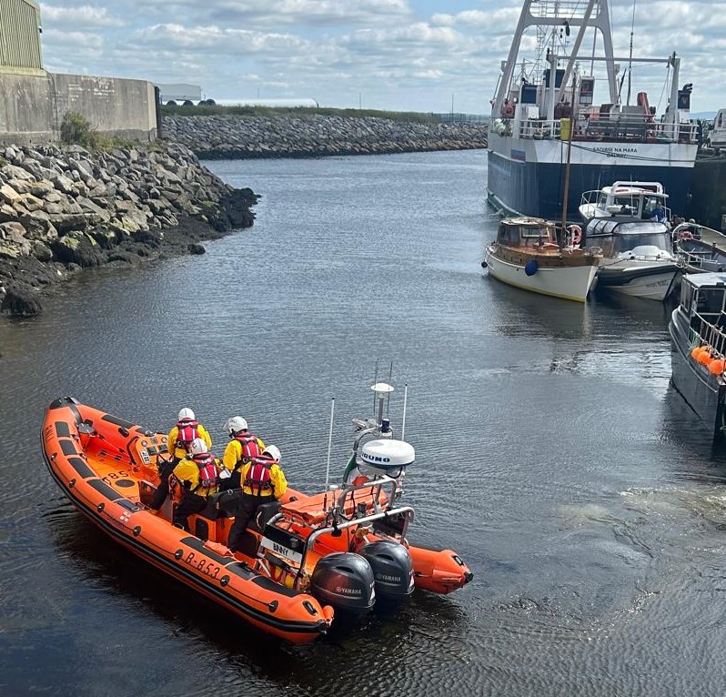 Inshore lifeboat with four crew on board leaving the pontoon at Galway Lifeboat Station after launching. Low tide has exposed rocks on the channel to left of the lifeboat and there are a number of fishing and other boats tied up on the right had side of the channel.