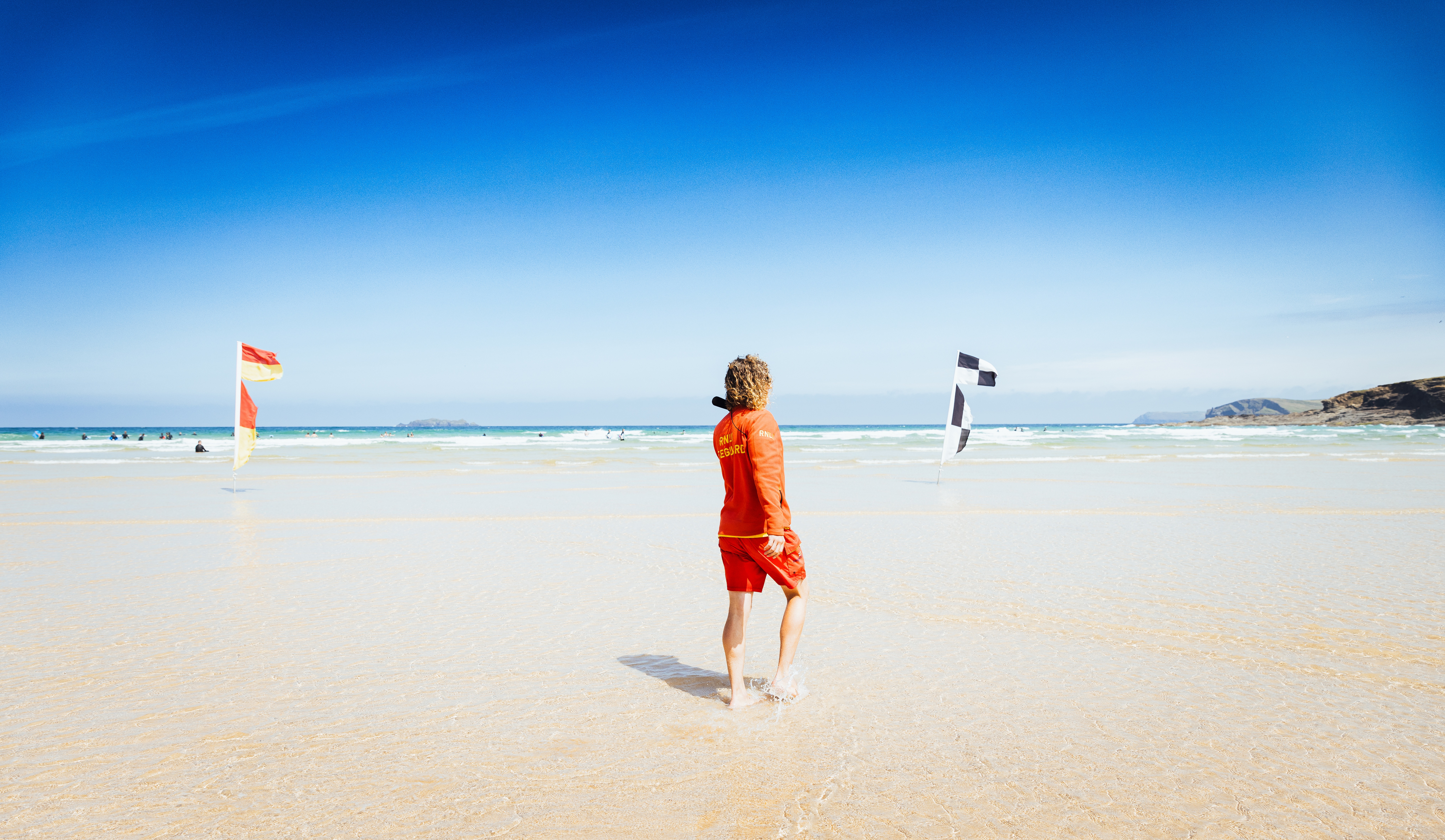 RNLI lifeguard stands between safety flags on sunny beach.
