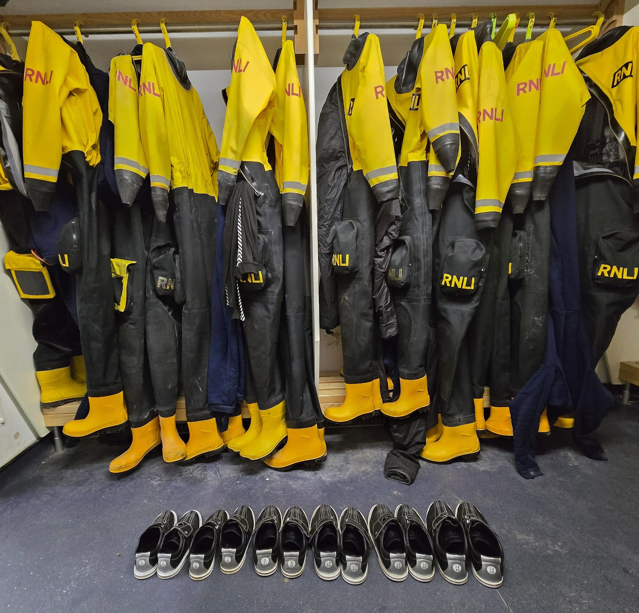 New Brighton RNLI's kit room. Visible are a rack of dry suits, some missing, currently in use. On the floor in front is a neat row of identical bowling shoes.
