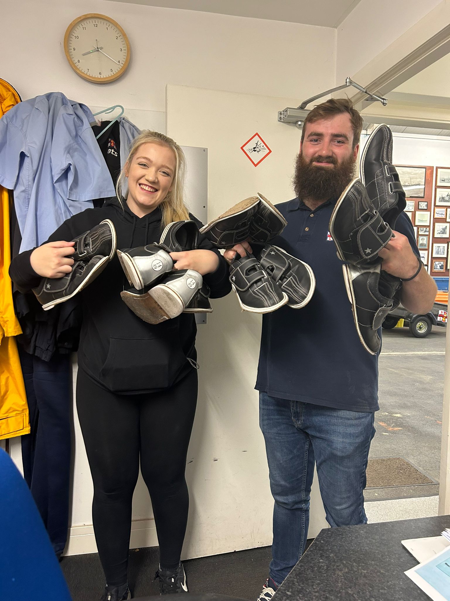 Volunteers Tom and Emily stand in an office, each holding many bowling shoes in their hands!
