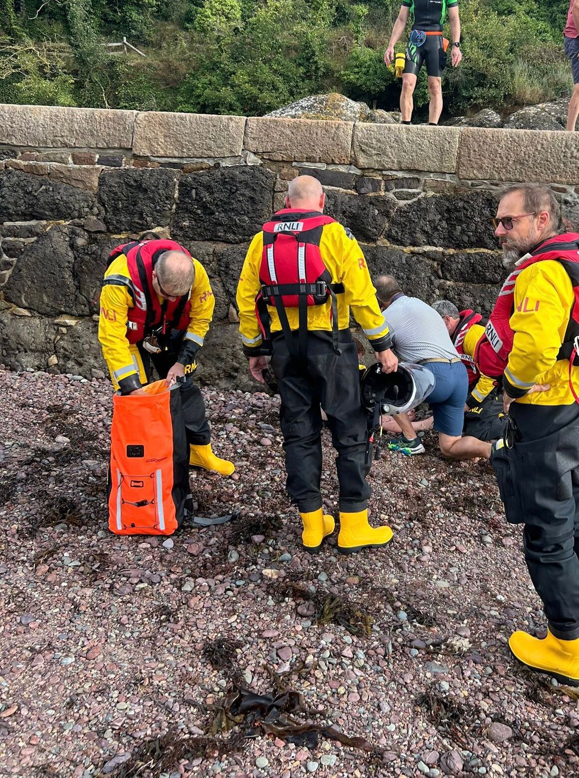 RNLI Jersey crew, kitted up in their dry suits, provide casualty care to an injured boy, who had fallen off the slip, to the side of the slip at Little St Catherine's
