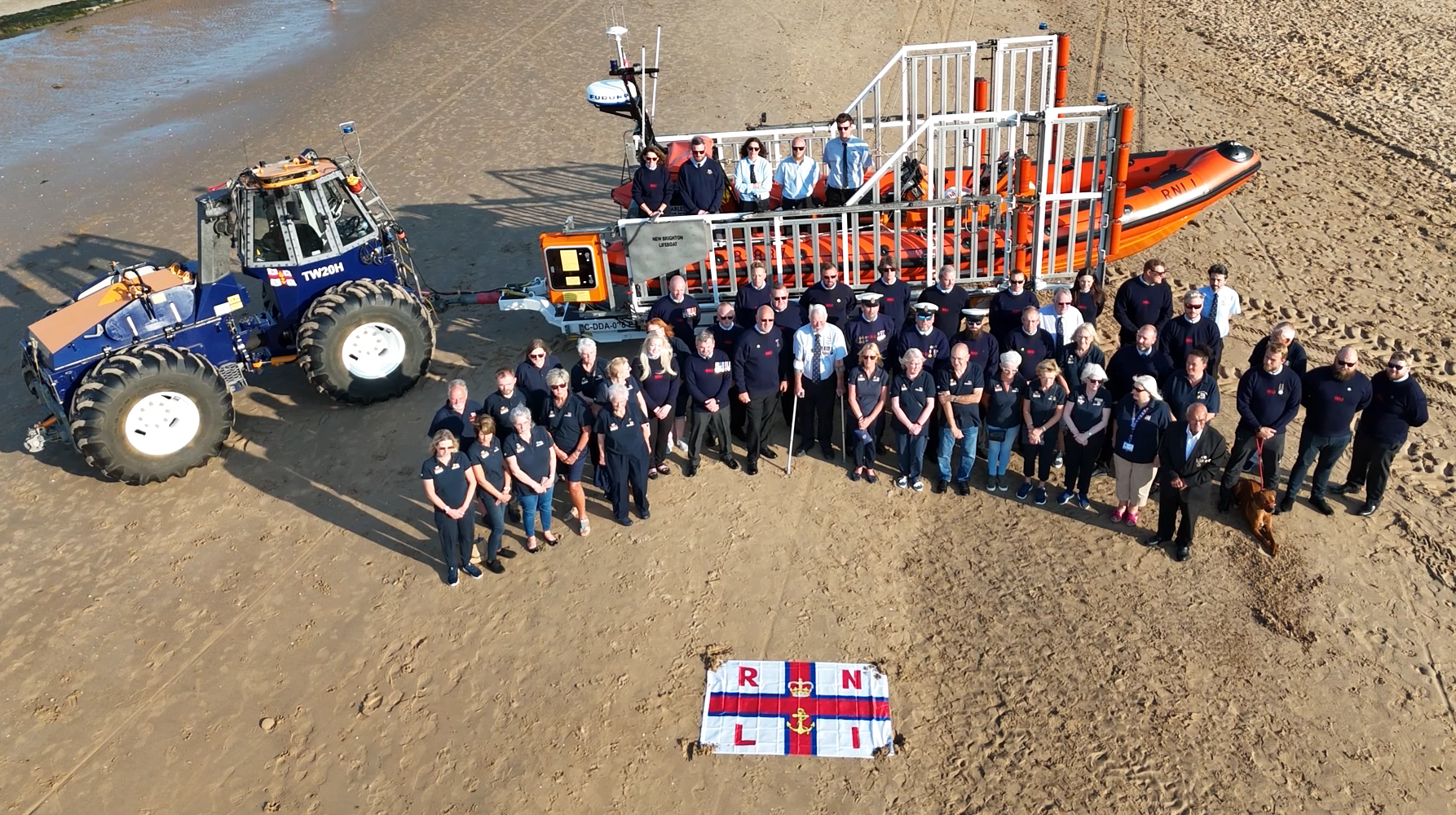 New Brighton RNLI's volunteers are gathered on the beach, with tractor and boat behind them, and RNLI flag in front, posing for a photo taken from a drone.