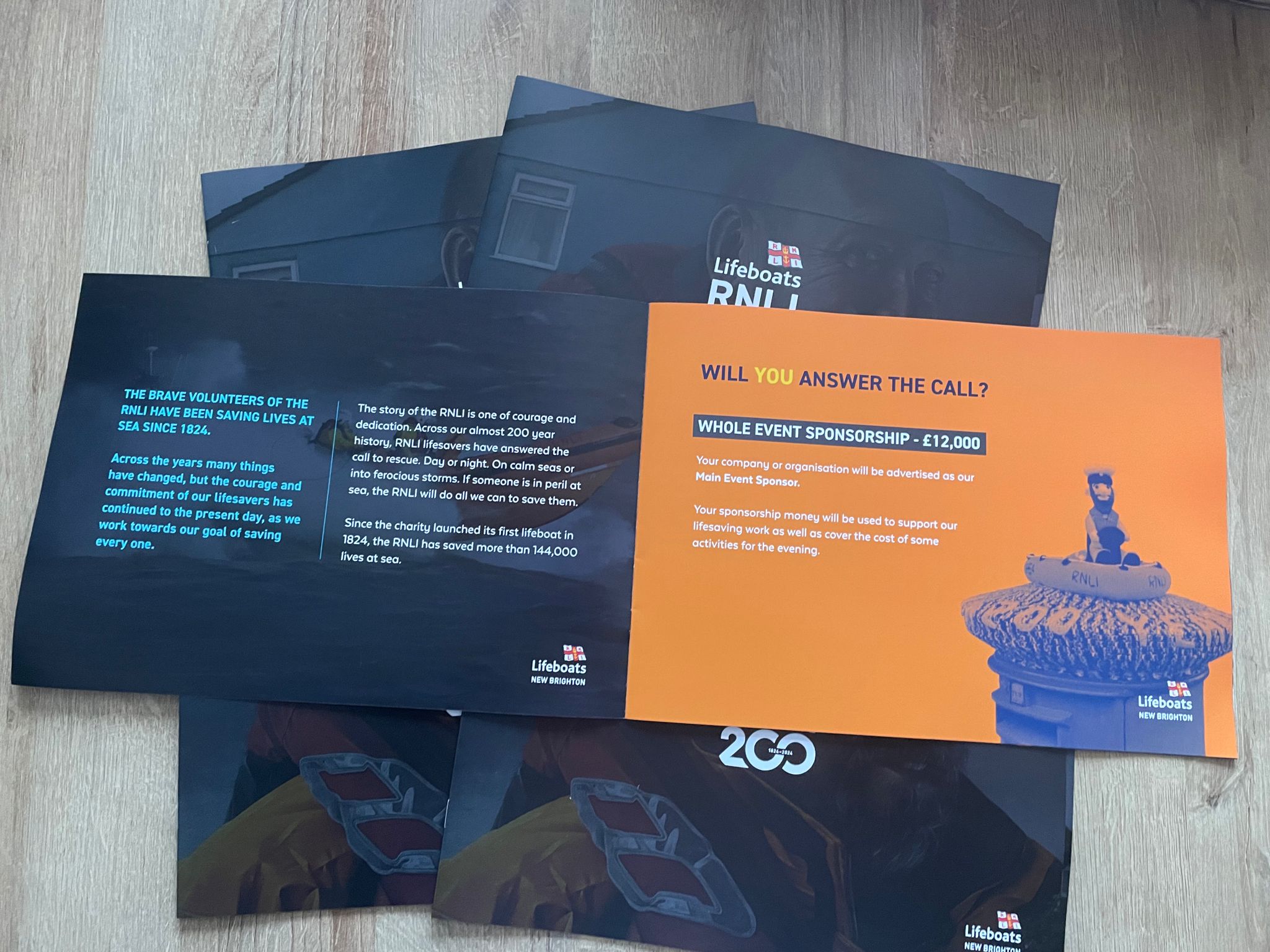 One of the sponsorship booklets lies open against a pile of others on a table. The left page is dark blue with white text, the right page is bright orange with blue text and imagery.