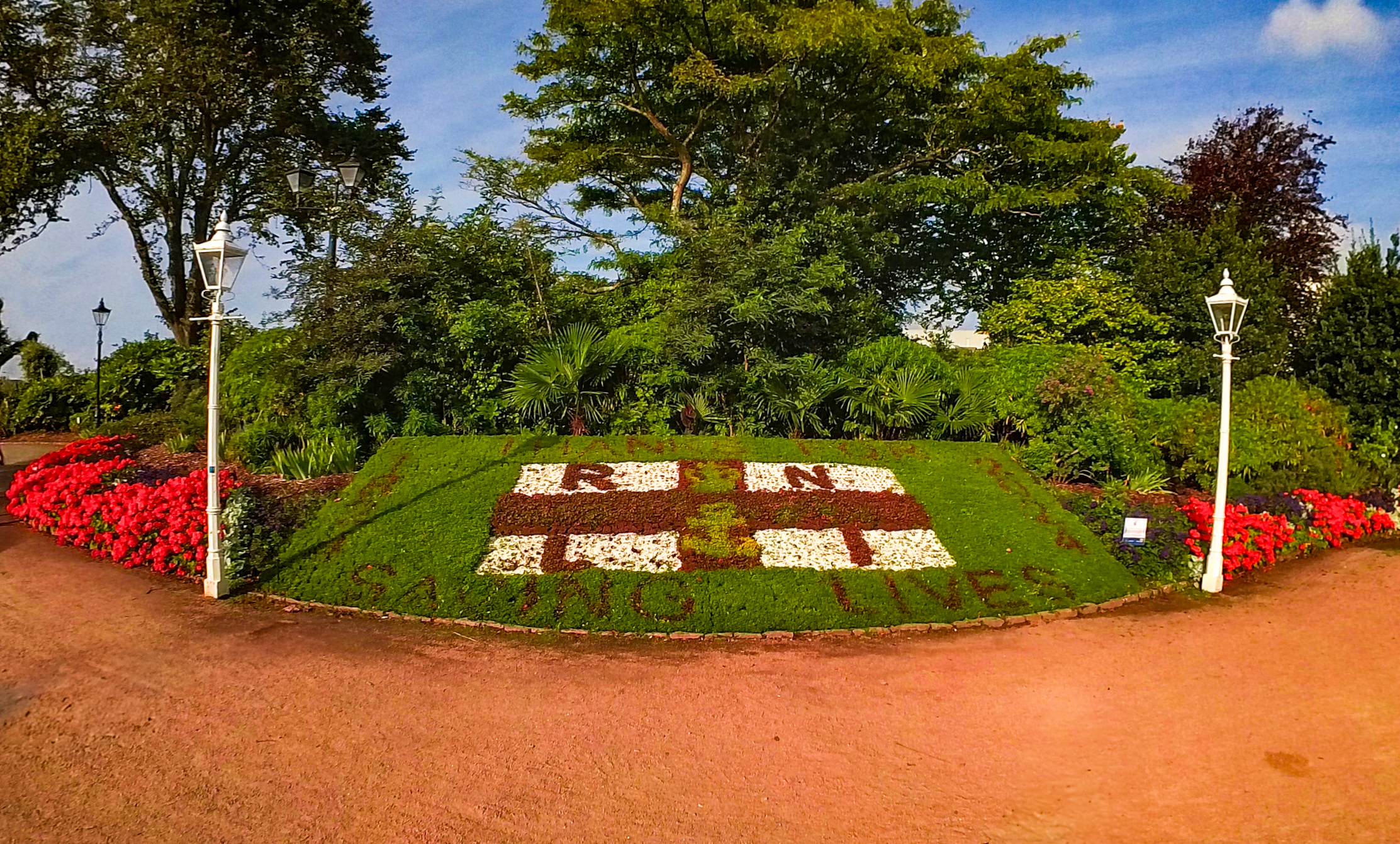 A wide angle shot of the decorative flowerbed at Howard Davis Park with the gravel path in the foreground and the flowerbed in the background under a blue sky and mature trees. The flowerbed is the RNLI flag with the words 'Thanks for' and 'Saving lives' at the top and bottom of the flag and '1824' to the left and '2024' to the right.