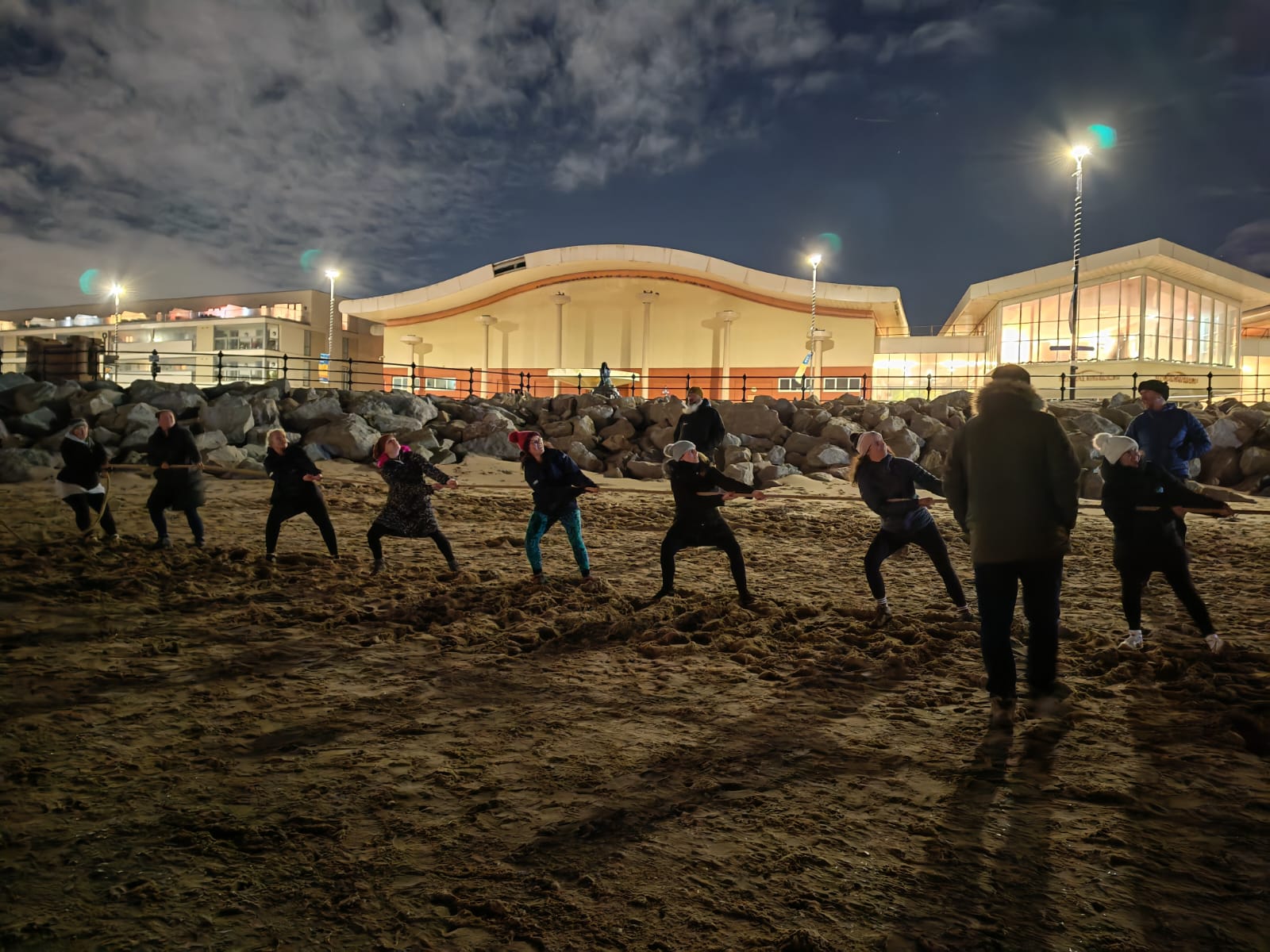 A night time shot, on New Brighton beach. Floral Pavilion theatre visible in the background, whilst on the beach in the foreground, a team of volunteers from New Brighton RNLI hold and pull hard on a long rope. Others stand nearby watching.