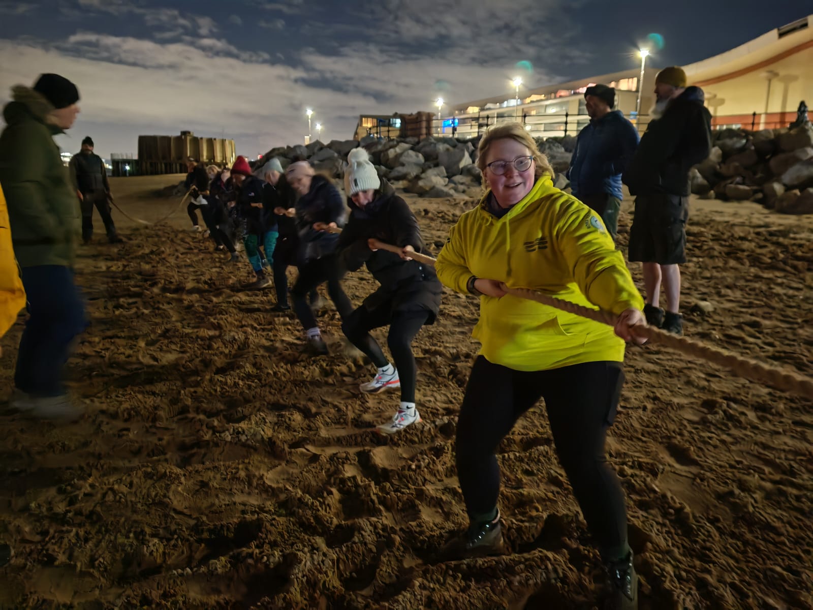 A night time shot, on New Brighton beach. On the beach in the foreground, a team of volunteers from New Brighton RNLI hold and pull hard on a long rope. Others stand nearby watching.
