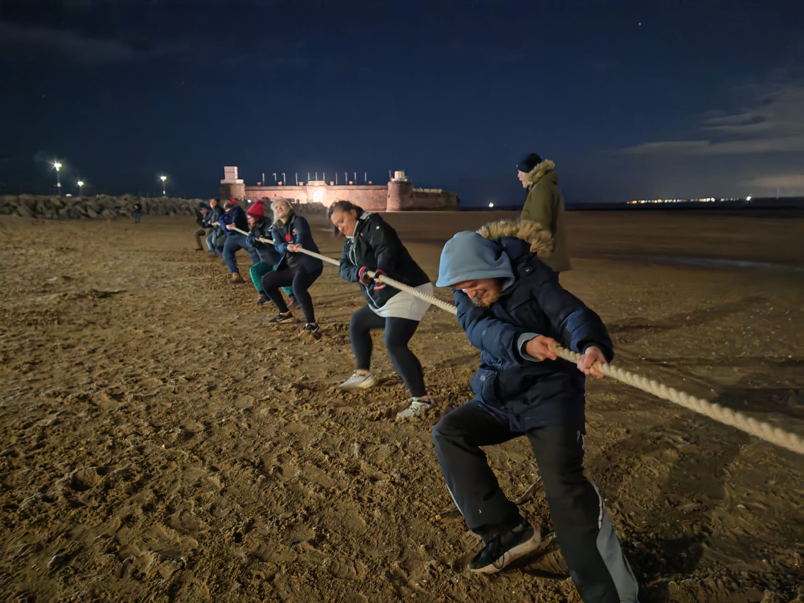 A night time shot, on New Brighton beach. On the beach in the foreground, a team of volunteers from New Brighton RNLI hold and pull hard on a long rope. Others stand nearby watching.