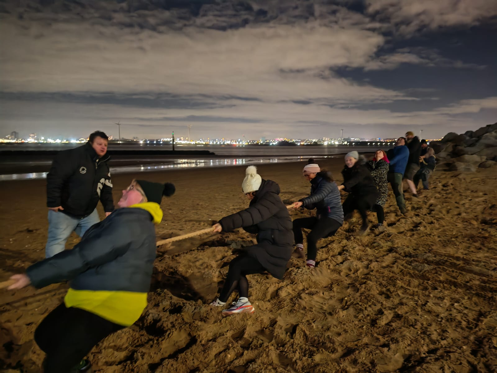 A night time shot, on New Brighton beach. On the beach in the foreground, a team of volunteers from New Brighton RNLI hold and pull hard on a long rope. Others stand nearby watching.