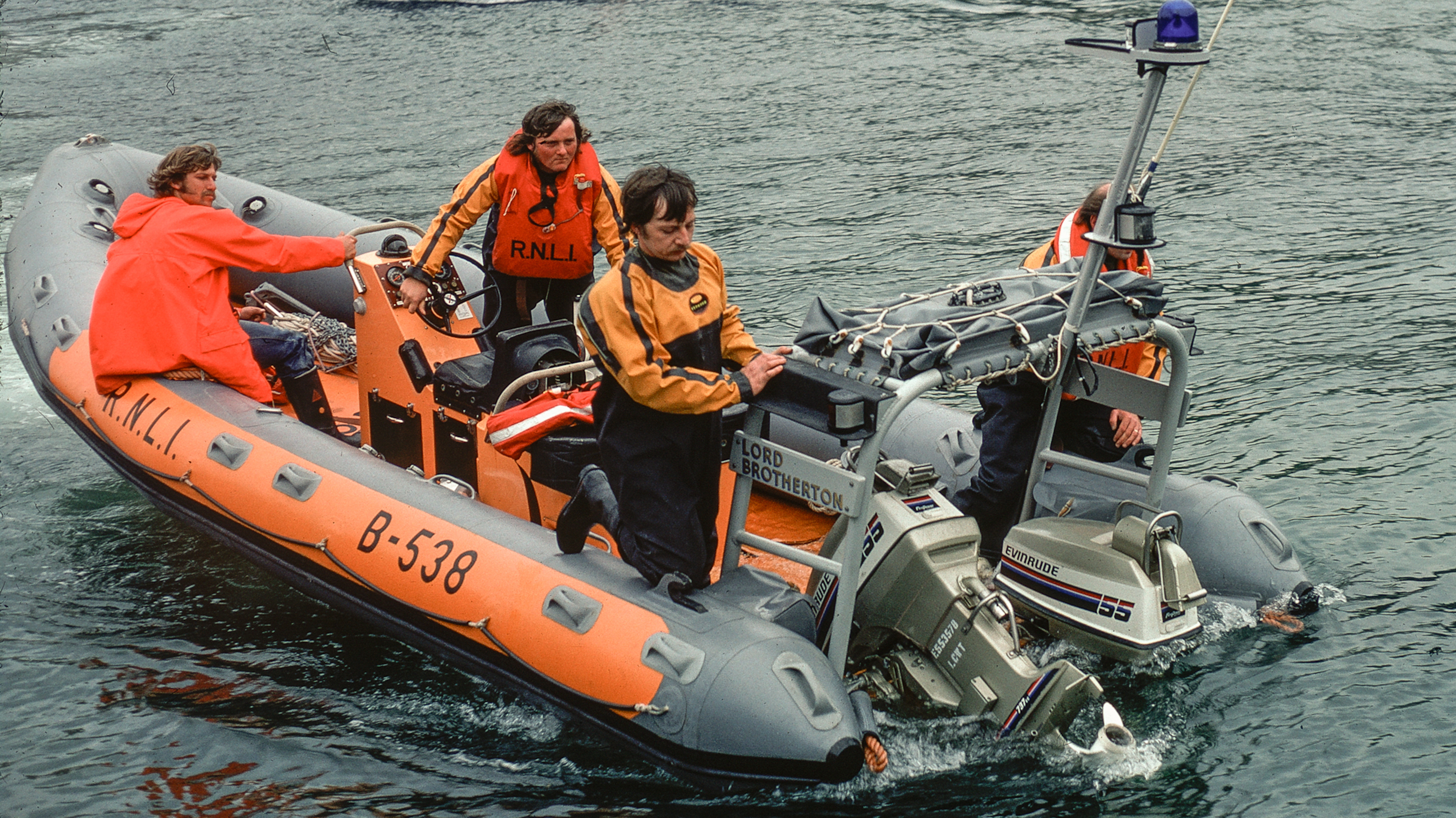 An orange and grey inflatable RNLI lifeboat, 'Lord Brotherton' (B-538), returns to its station on calm water. Four volunteer crew members in waterproof gear manage the boat, one operating controls, and others securing the vessel.