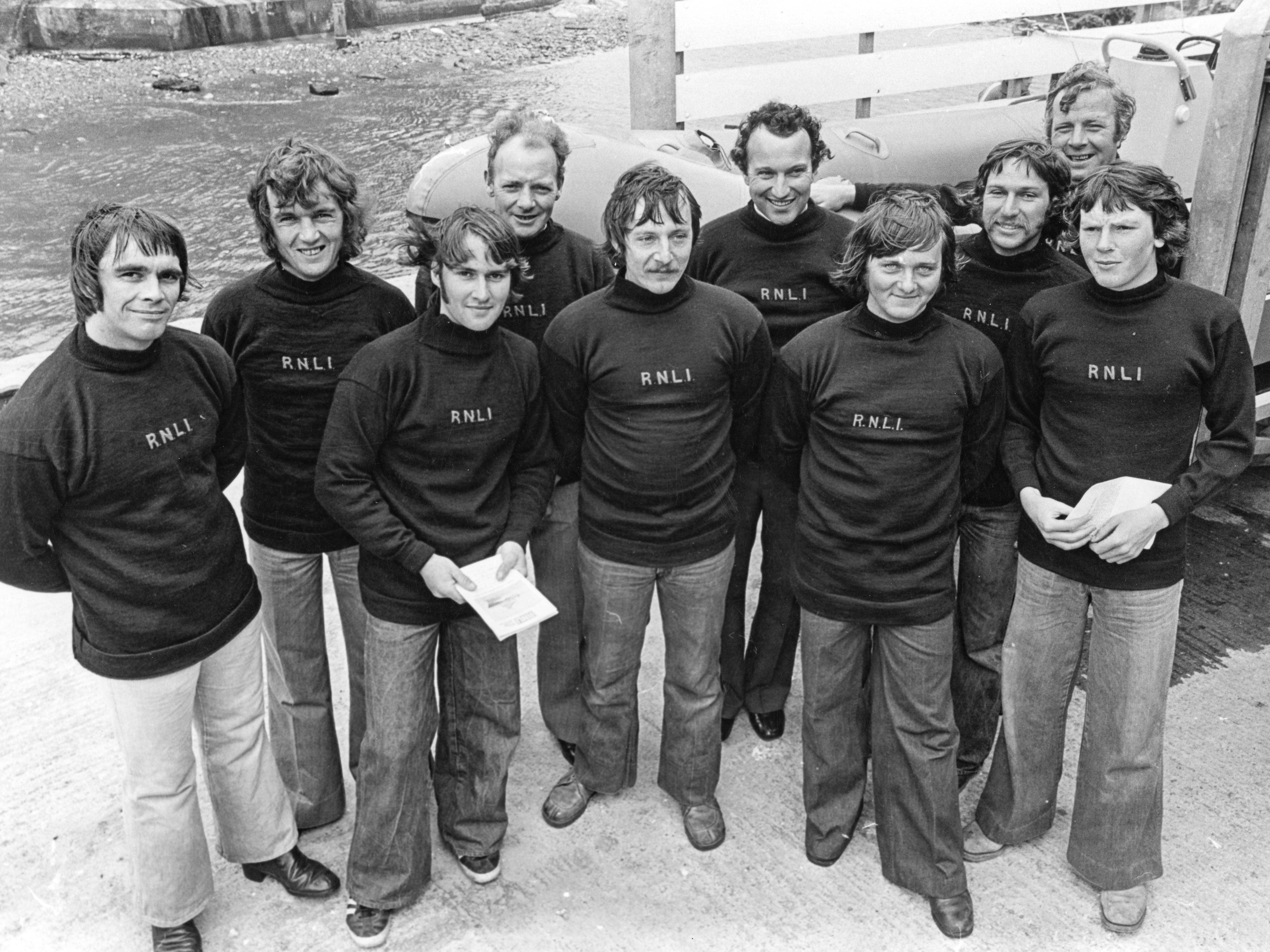 This black-and-white photograph shows a group of ten male RNLI (Royal National Lifeboat Institution) volunteers standing together, smiling, and posing for the camera. They are wearing matching RNLI sweaters and flared trousers, indicative of the late 1970s fashion. The group is outdoors, and behind them, part of the lifeboat B-538 Lord Brotherton is visible, along with a body of water and dock equipment. The photo was taken during the 1978 inauguration of the lifeboat.
