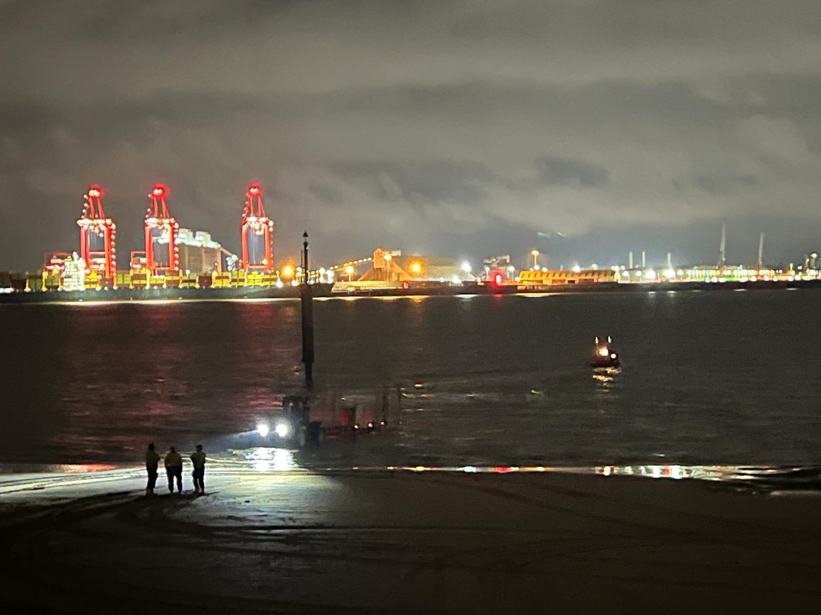 A wide shot of New Brighton beach at night, Liverpool skyline visible in the background. At the river's edge is the tractor, with boat just ahead having just launched. Crew stand and watch on from the beach.