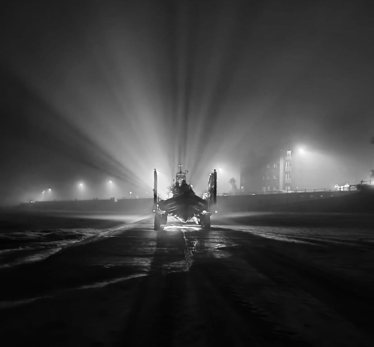 New Brighton RNLI's tractor, carrying the lifeboat, is heading toward the photographer, launching on service. In the fog, beams of light can be seen shining out from the lights behind into the sky. Black and white.